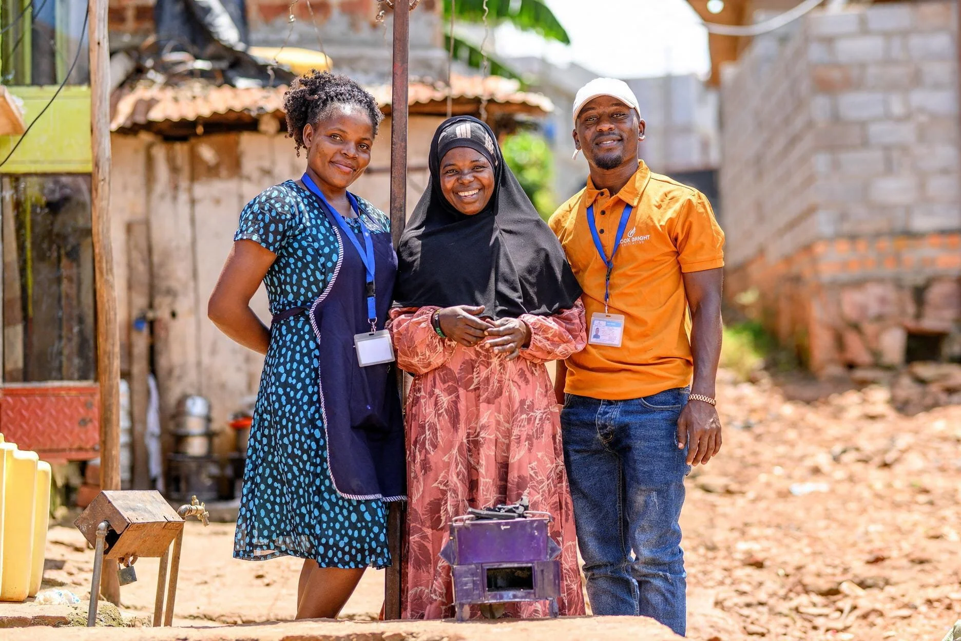 Three people smiling outdoors in a developing area, wearing ID badges, standing next to each other with an informal background of unfinished buildings and dirt ground.