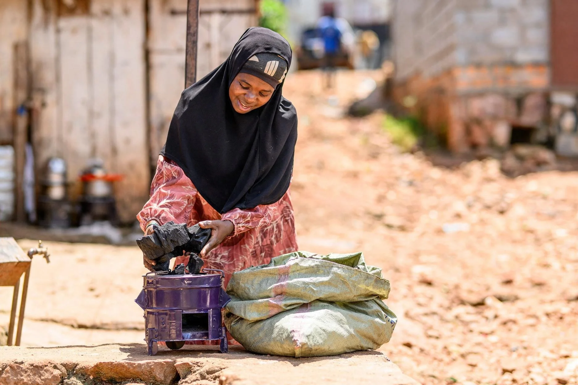 A woman in a black headscarf and patterned long-sleeve dress is gathering charcoal into a small purple cart outdoors with a large bag of charcoal beside her, in a rural setting.