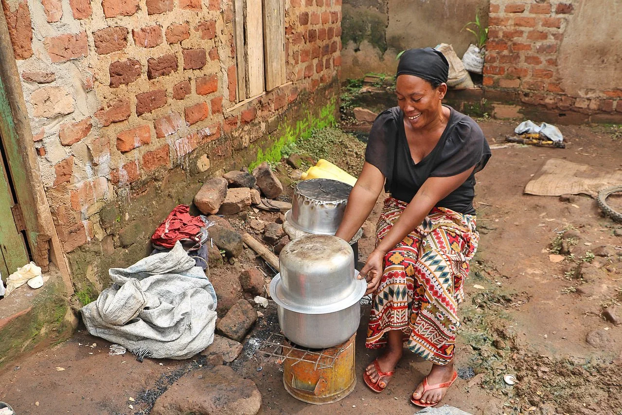 A woman is cooking outdoors using pots on an improved impact carbon stove inside a brick walled area.