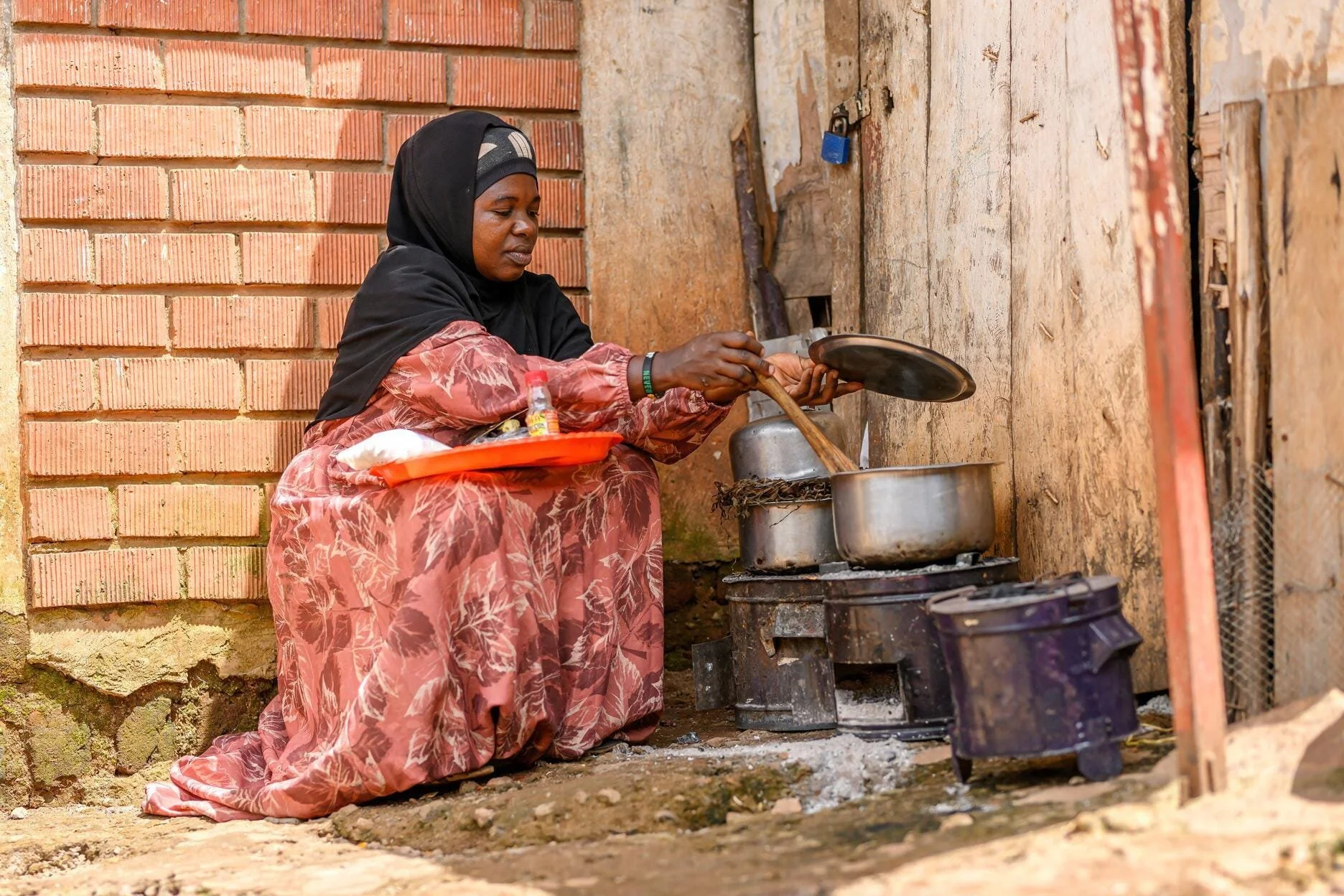 A woman wearing a headscarf and colorful dress is cooking outdoors on a small stove with two pots, surrounded by rustic walls and a rough ground.