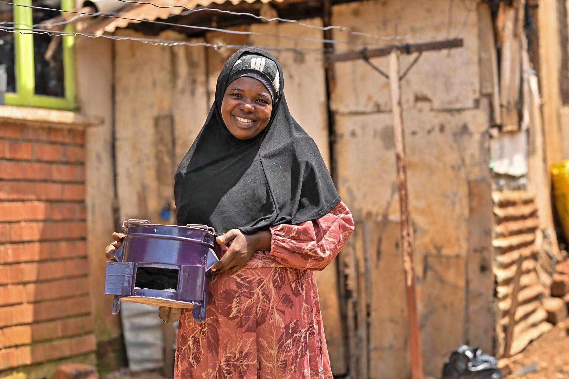 A smiling woman wearing a black hijab and a pink dress holding a purple and black container in an outdoor setting with a rustic background.