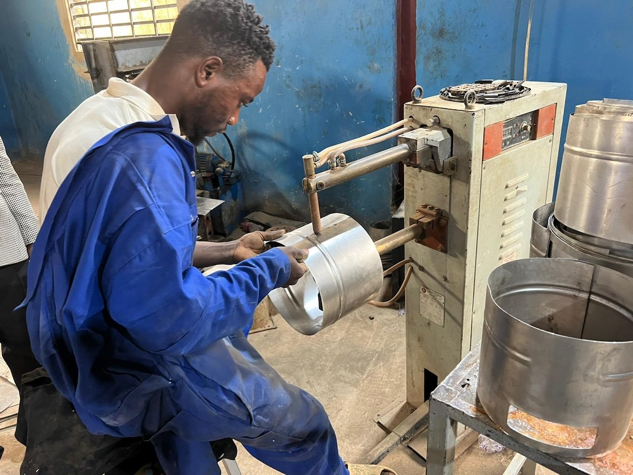 A man operating a metal can forming machine, shaping a metal can. He is wearing a blue jacket and working in a workshop environment.