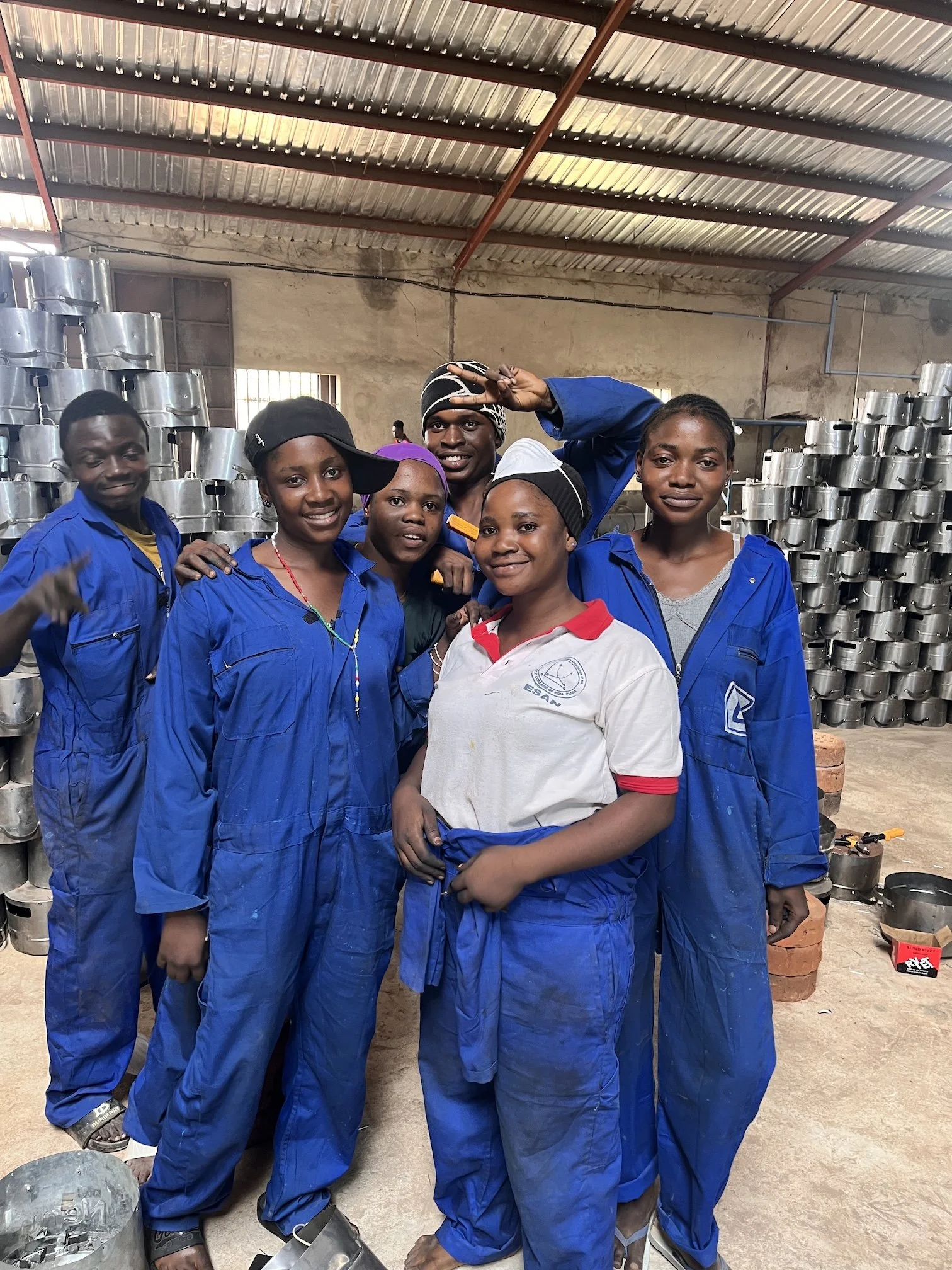 A group of six young people, four women and two men, in blue work uniforms, standing together inside a workshop with metal space heater parts stacked behind them. They are smiling and posing for the photo.