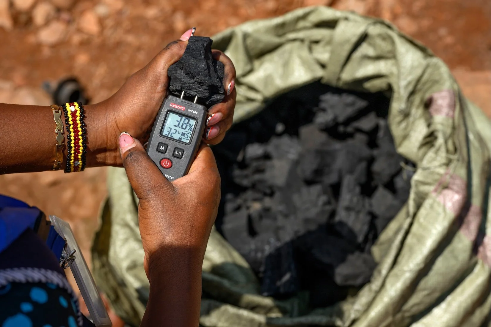 Person holding a moisture meter measuring black coal in a plastic bag, with a backpack filled with more coal behind.