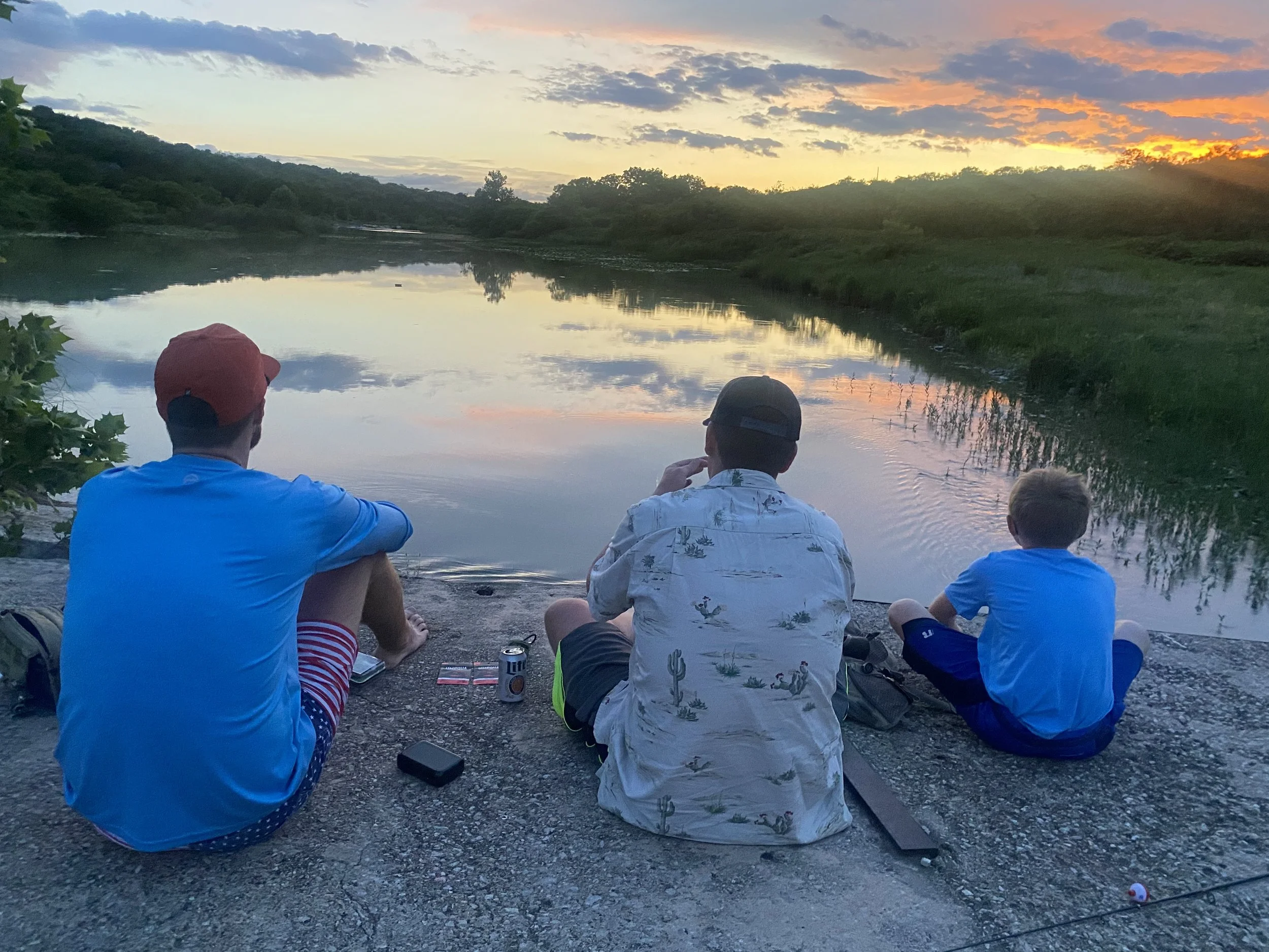 Three people sitting on a West Texas rocky riverbank, watching the sunset over a calm river, with trees and cloudy sky in the background.
