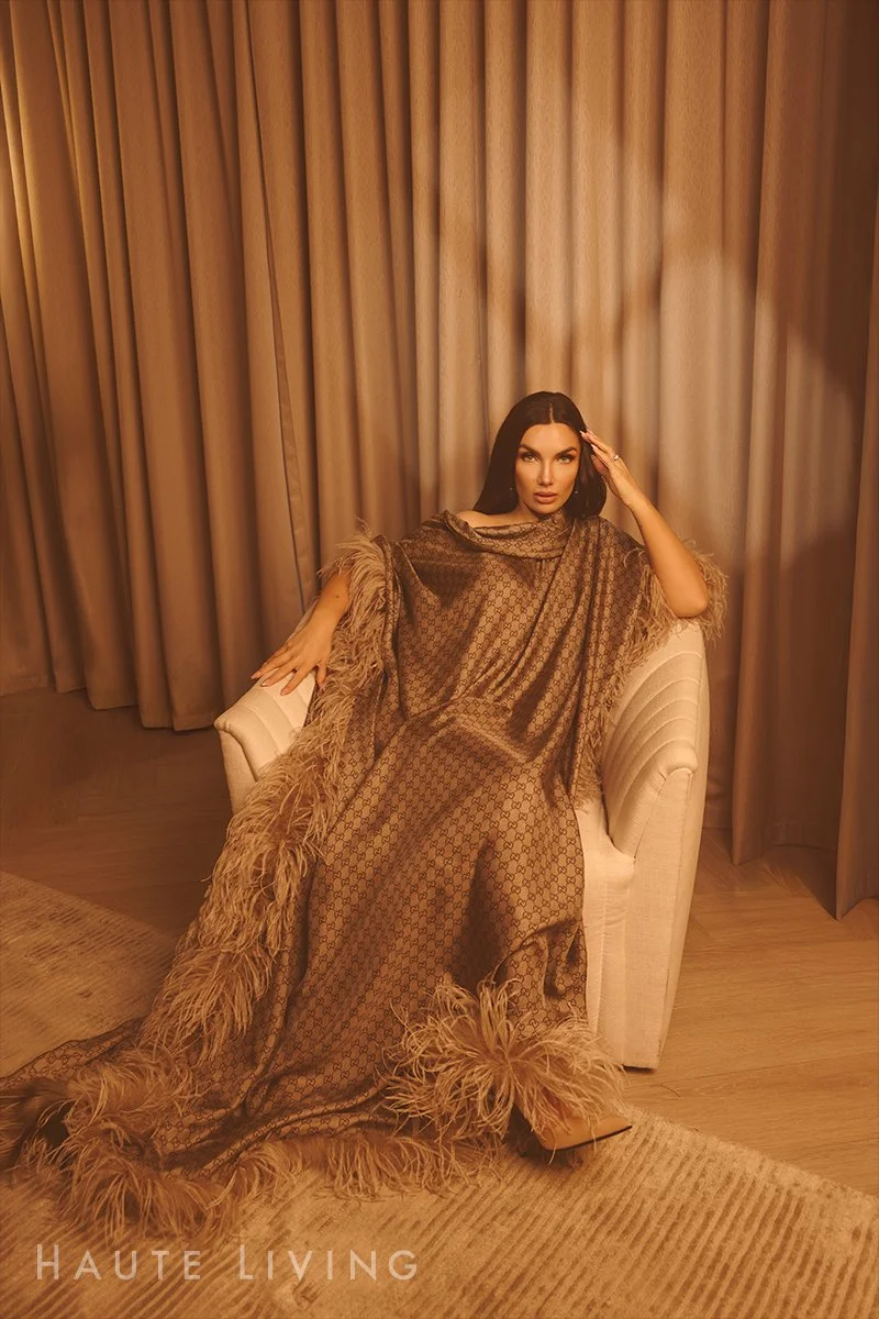 Elettra Lamborghini seated on a beige armchair in a room with beige curtains, wearing a patterned dress with feathered trim from Gucci by Demna.