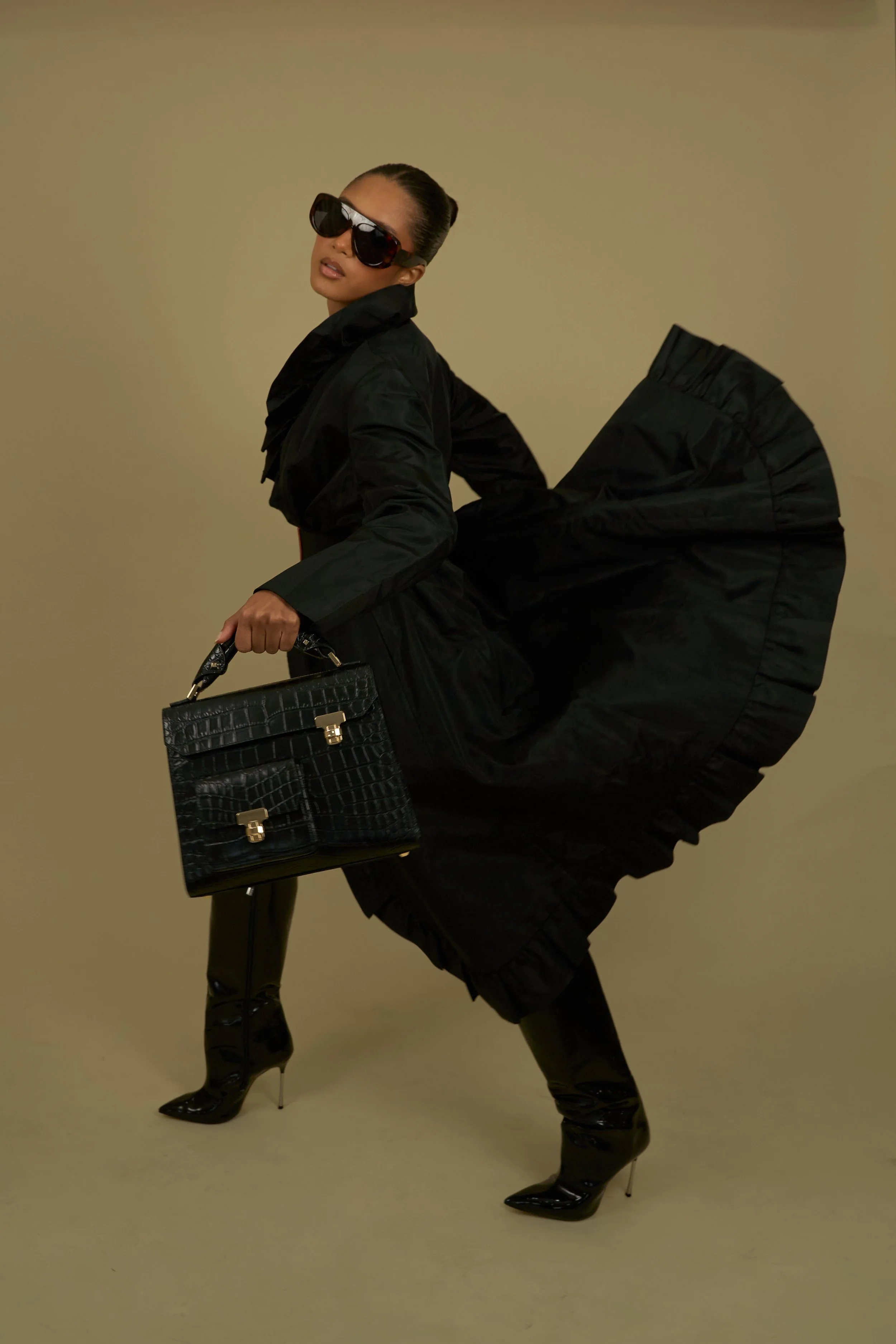 Fashion model wearing black oversized sunglasses, black puffer coat, black high-heeled boots, holding a black handbag, with a beige background.
