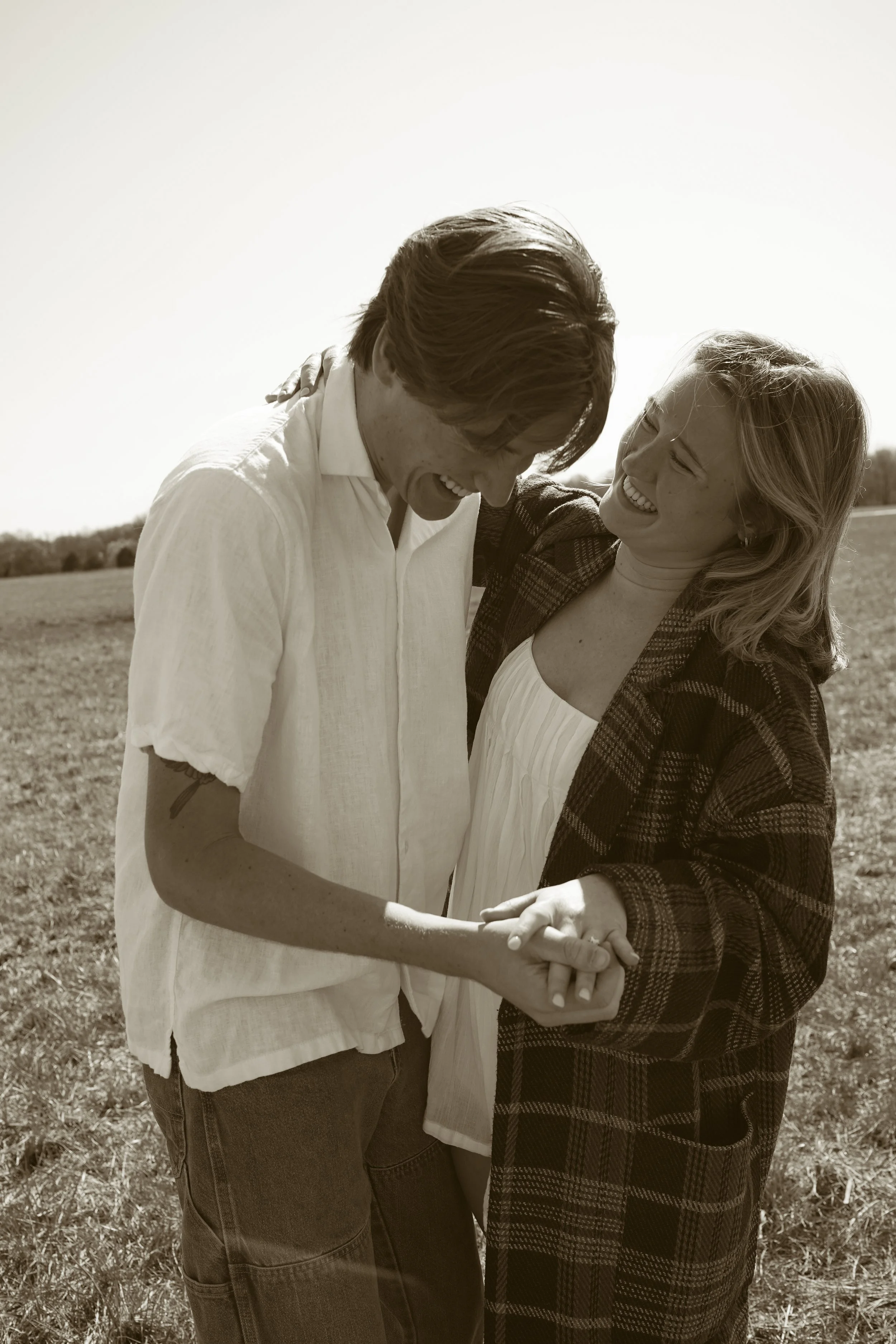 A couple smiling and holding hands in an open field on a sunny day, sharing a joyful moment.