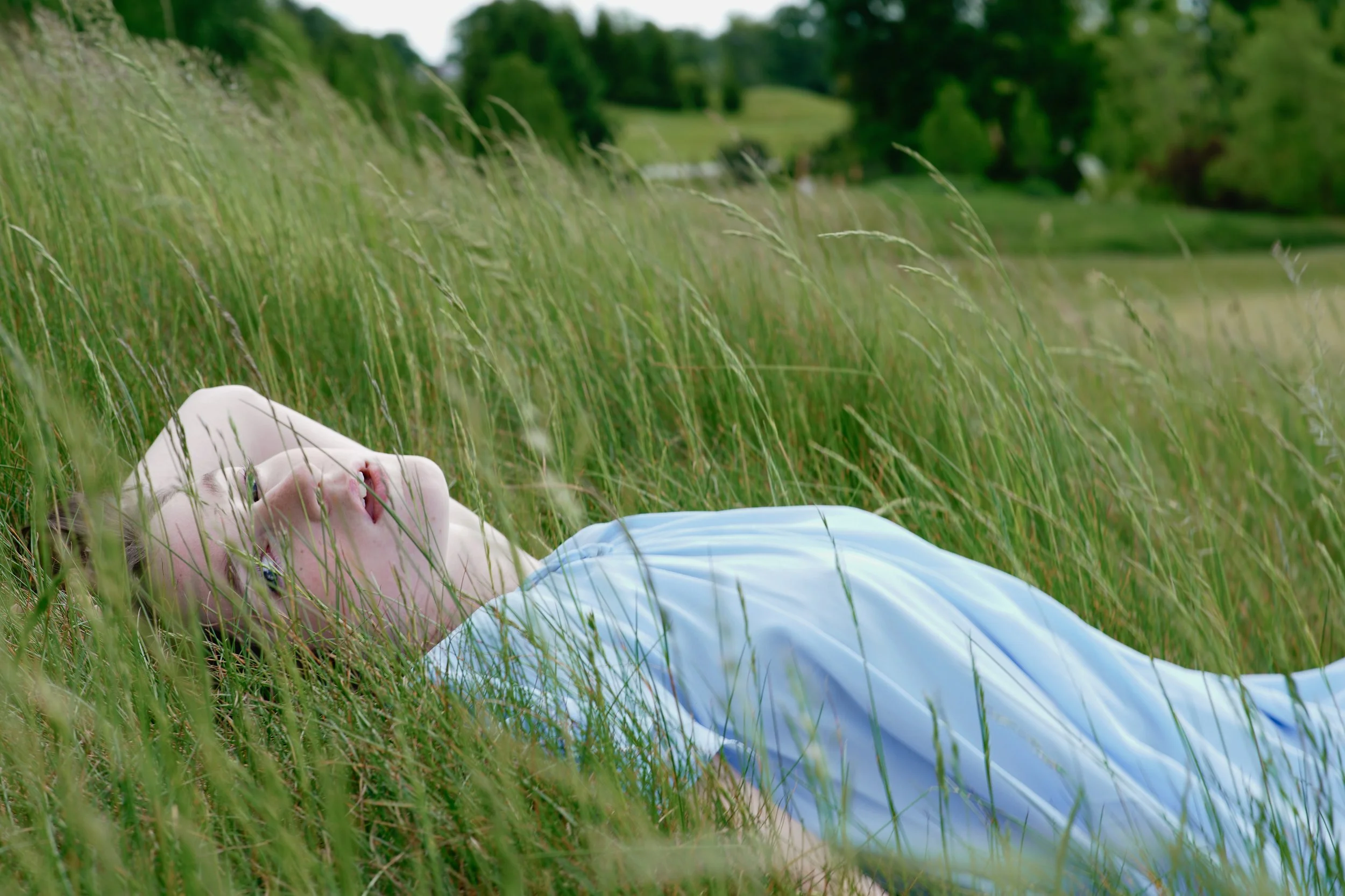 A person lying on the grass in a field with tall green grass, with trees and a cloudy sky in the background.