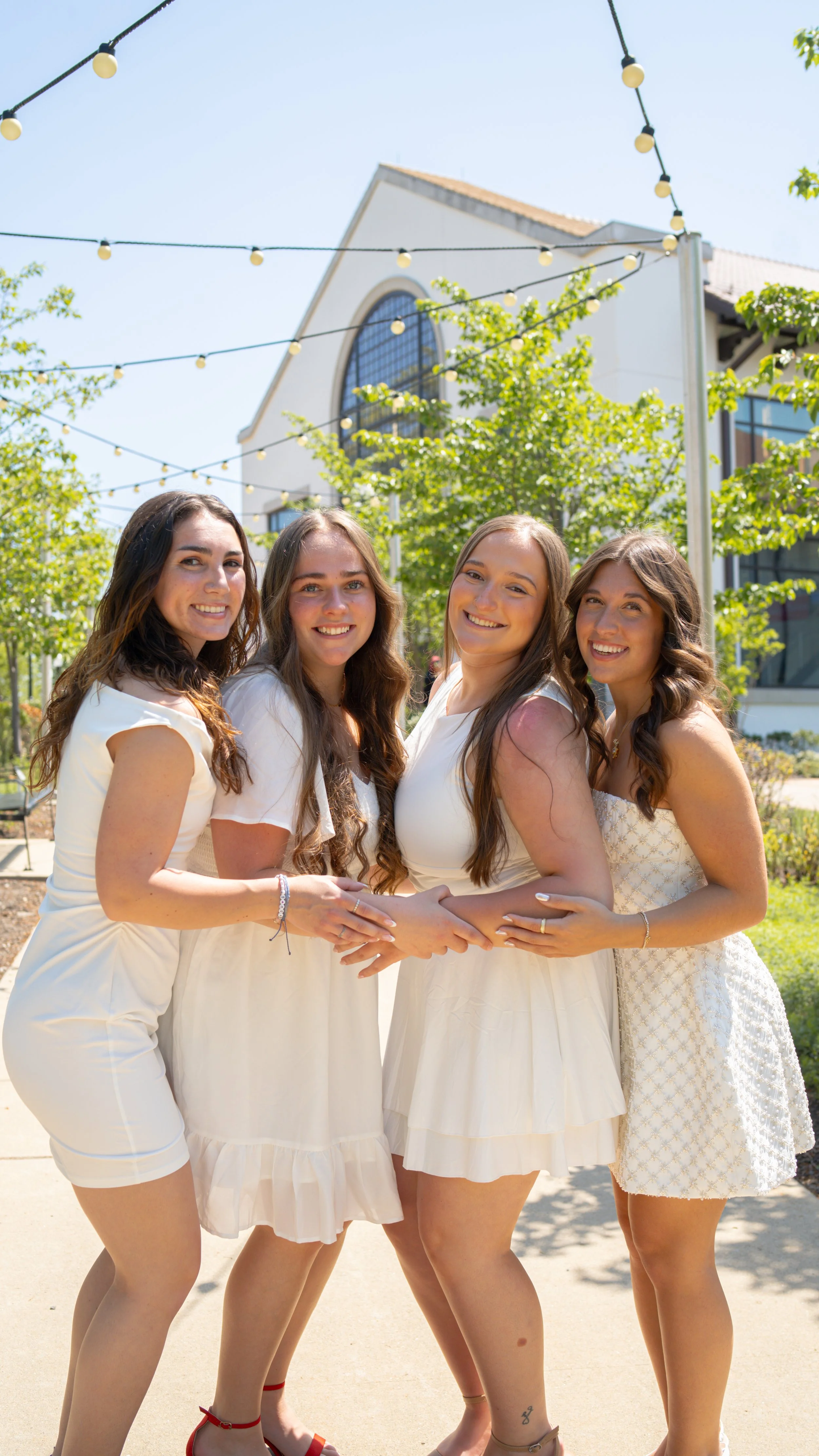 Four women dressed in white, smiling and hugging outdoors on a sunny day with string lights hanging above, green trees, a building with arched windows, and a clear blue sky in the background.