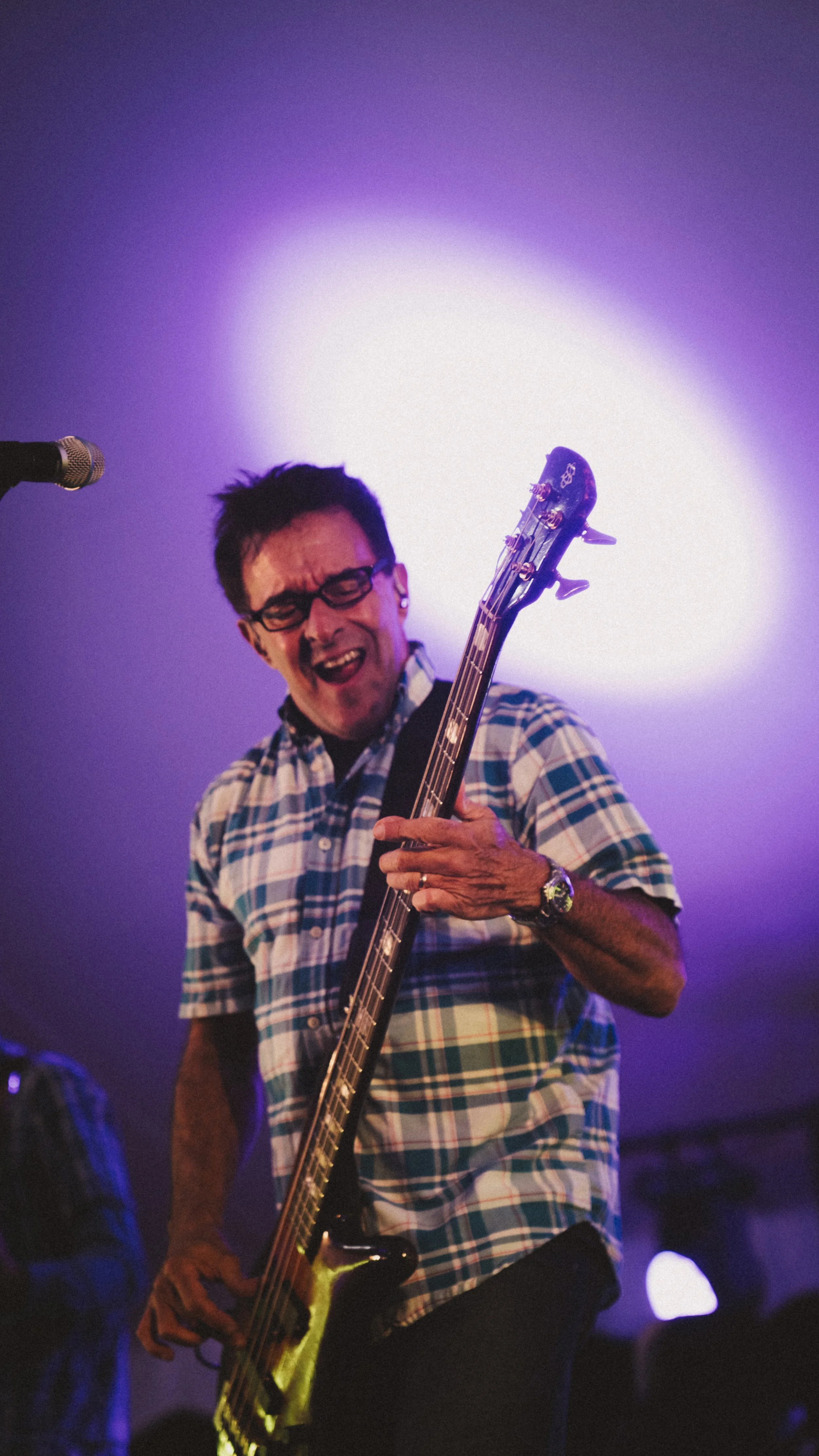 Man playing electric bass guitar on stage with purple and white lighting.