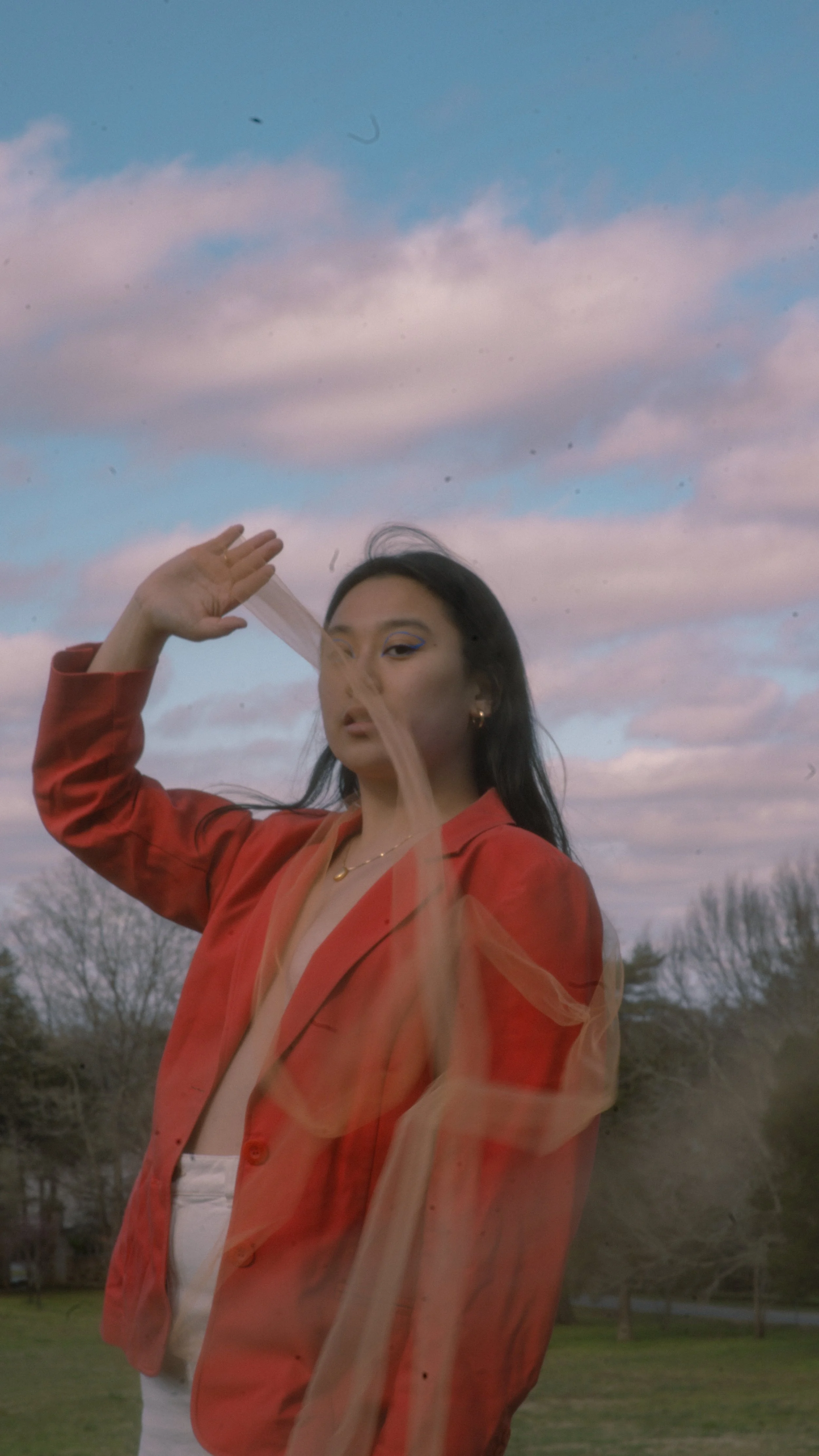 A woman with dark hair and makeup wearing a red jacket with sheer orange fabric outer layer, standing outdoors during sunset or sunrise, with cloudy sky and trees in the background.
