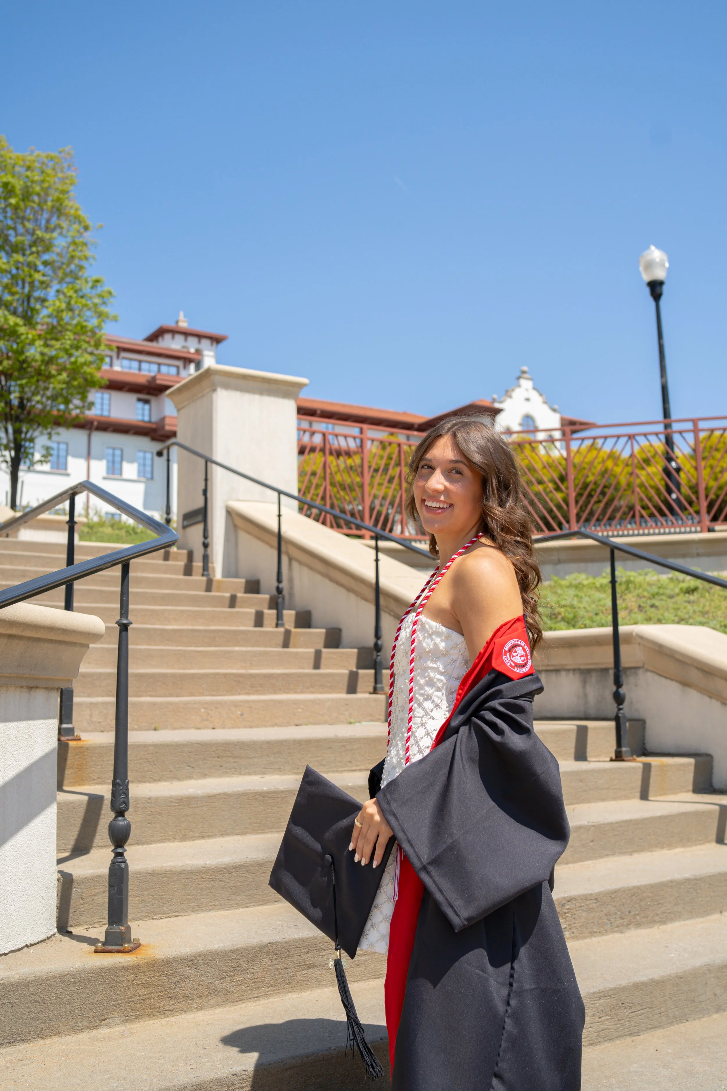 Female graduate in cap and gown smiling on outdoor stairs with blue sky and historic building in background.
