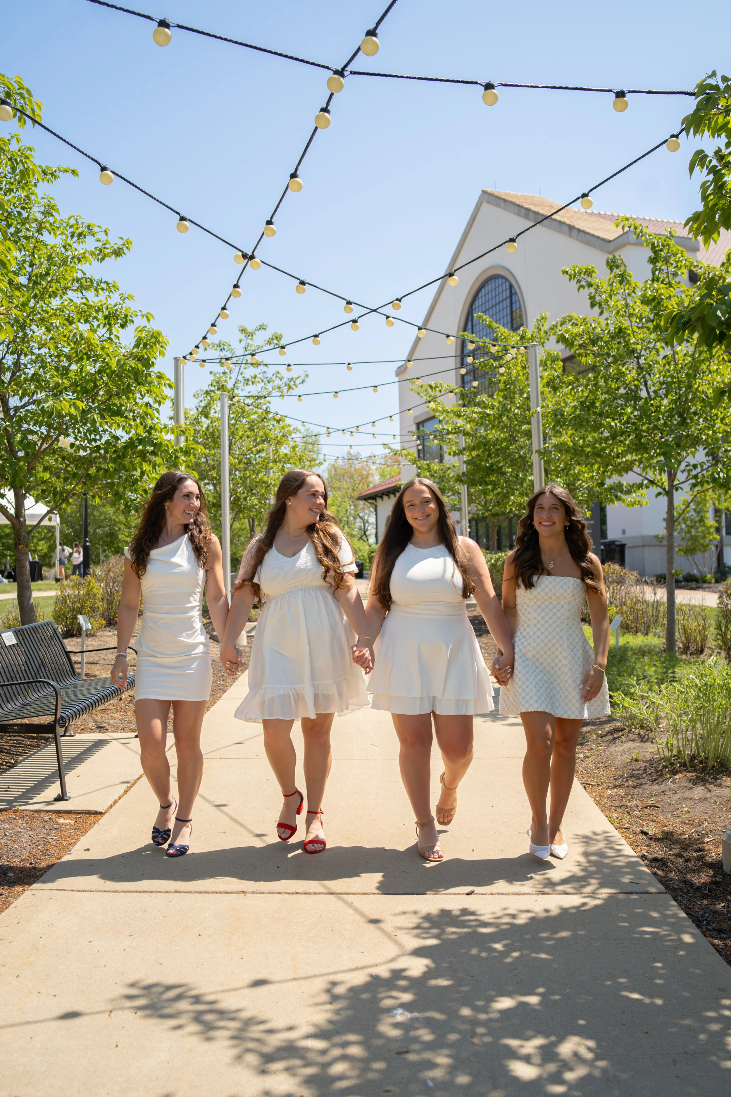 Four women dressed in white dresses walking hand in hand on a sunny sidewalk, with string lights overhead, trees on either side, and a building with large arched windows in the background.
