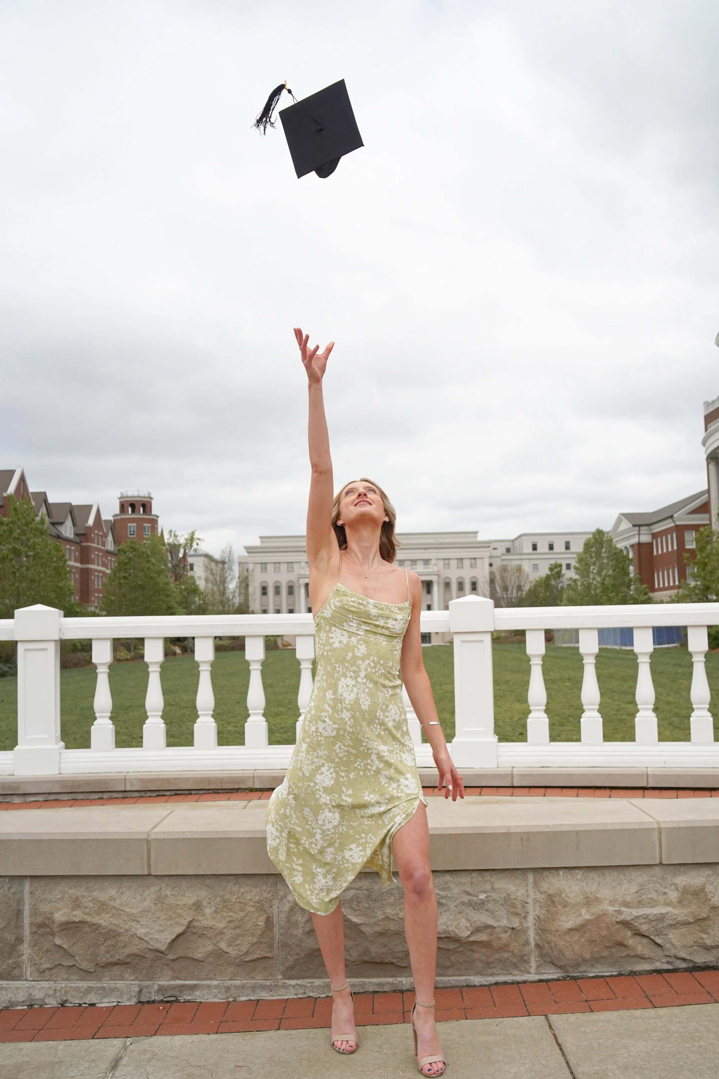 Woman in a yellow floral dress and high heels standing outside on a stone platform, tossing a black graduation cap into the air under cloudy skies, with buildings and greenery in the background.