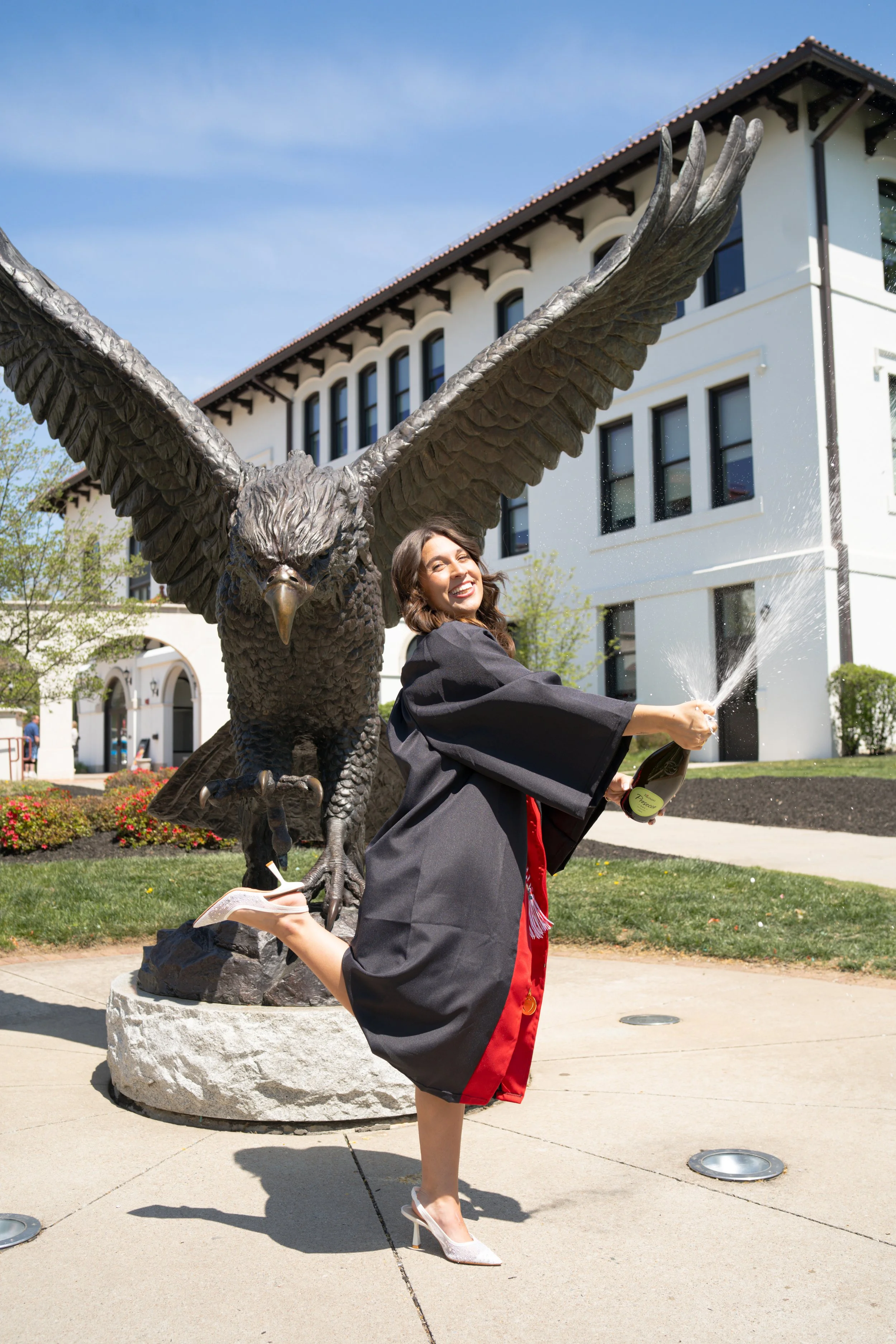 Woman in a graduation gown and heels wearing a red stole, celebrating outdoors in front of a large eagle statue by spraying champagne.