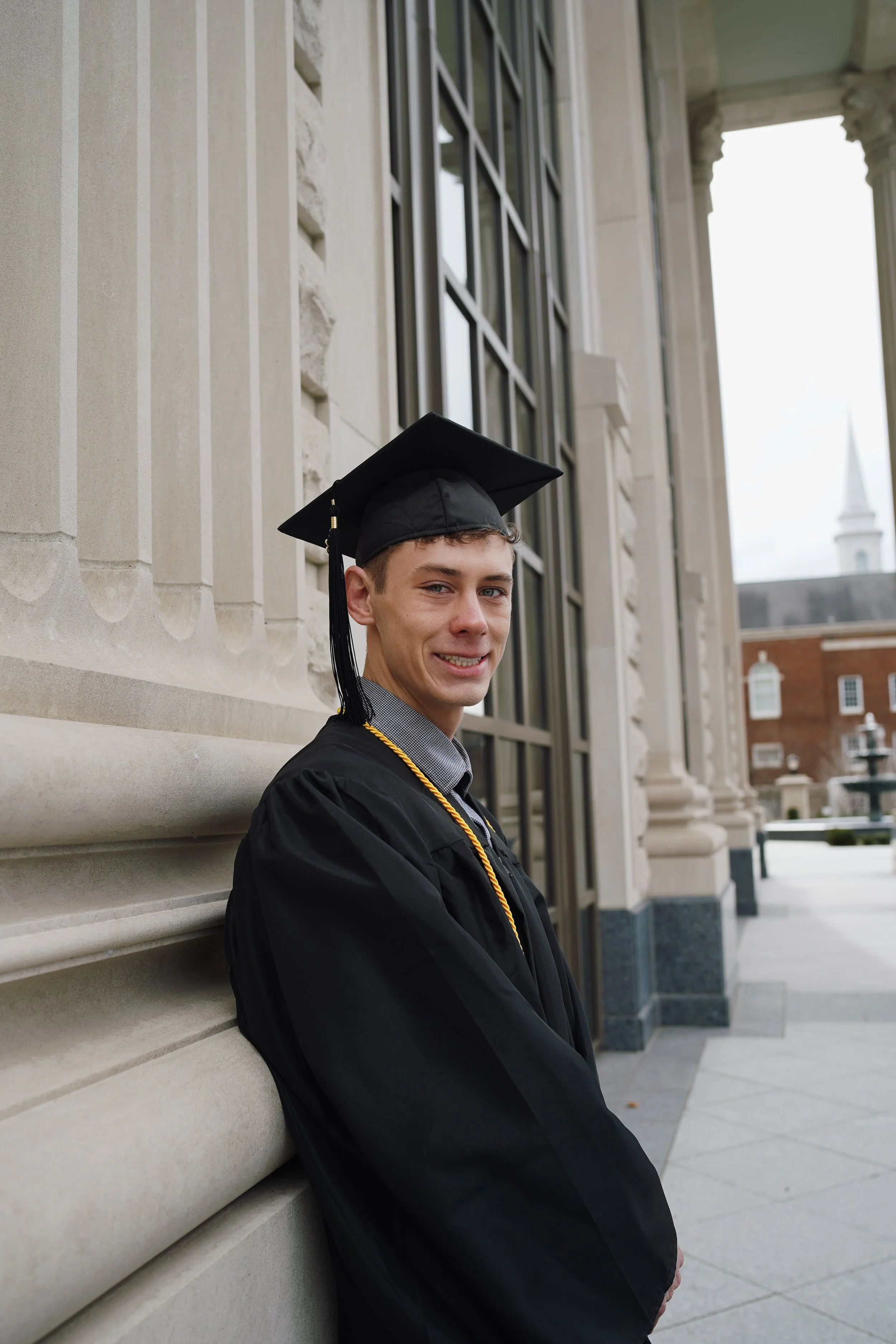 Young man in graduation cap and gown leaning against a stone building outside.