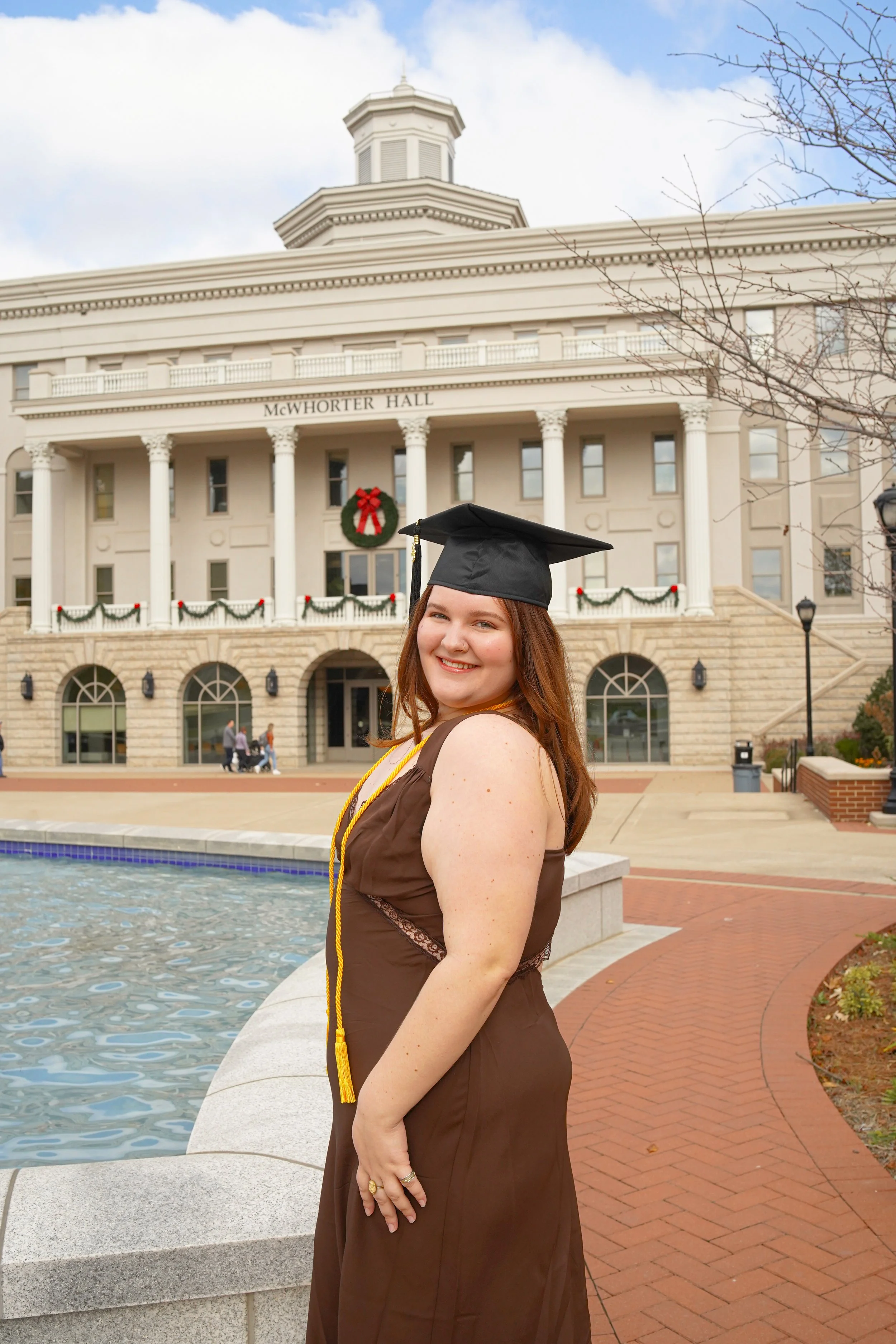 A young woman in a brown graduation gown and black cap with a yellow cord, smiling as she poses outdoors in front of McWhorter Hall decorated with Christmas wreaths and garlands, near a fountain and brick walkway, with a partly cloudy sky.