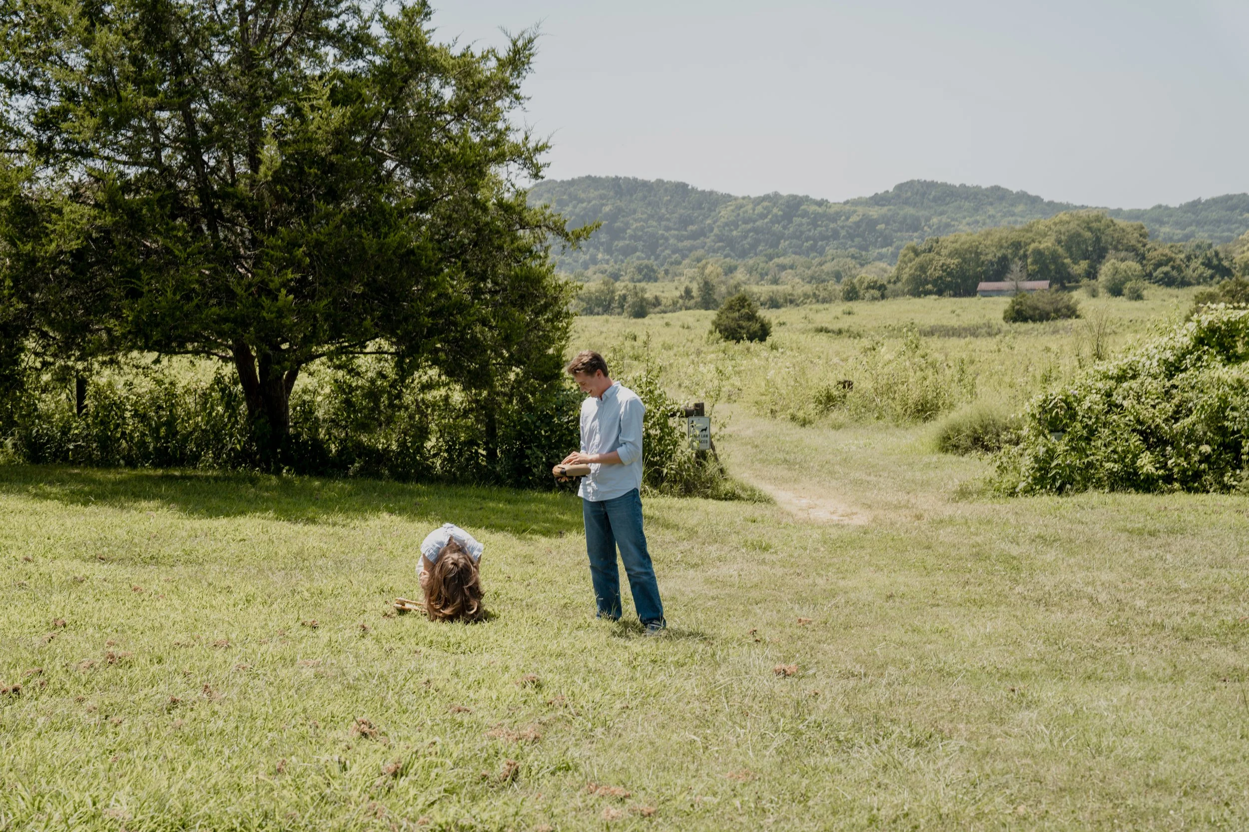 A young man and a girl with long hair are outdoors in a grassy field with green hills in the background. The young man is wearing a white shirt and blue jeans, and the girl is sitting on the ground. There are trees, bushes, and a small white sign nea