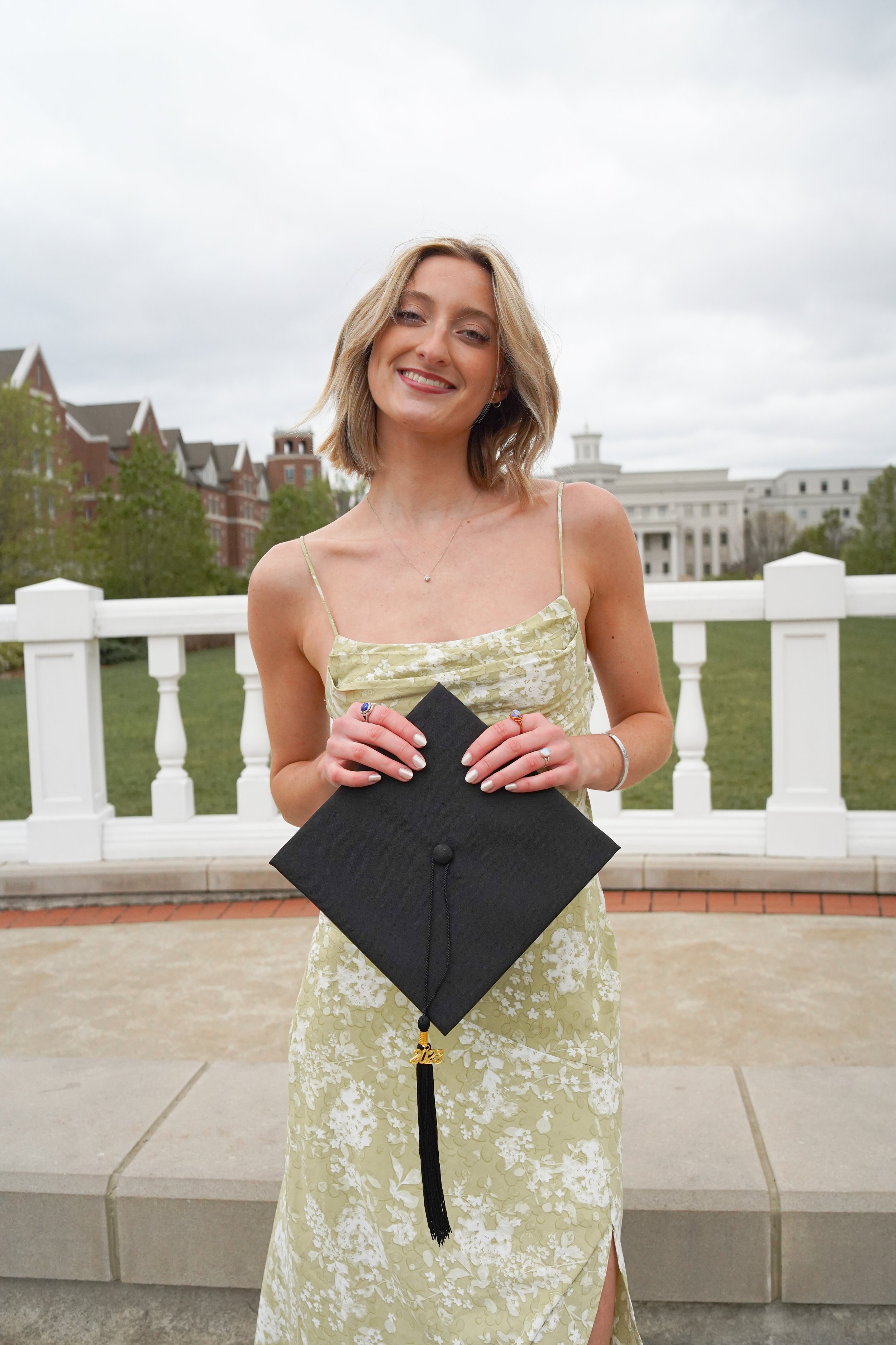 A young woman in a yellow floral dress holds a black graduation cap with a "2023" tassel, standing outdoors on a cloudy day in front of a white fence with historic buildings in the background.