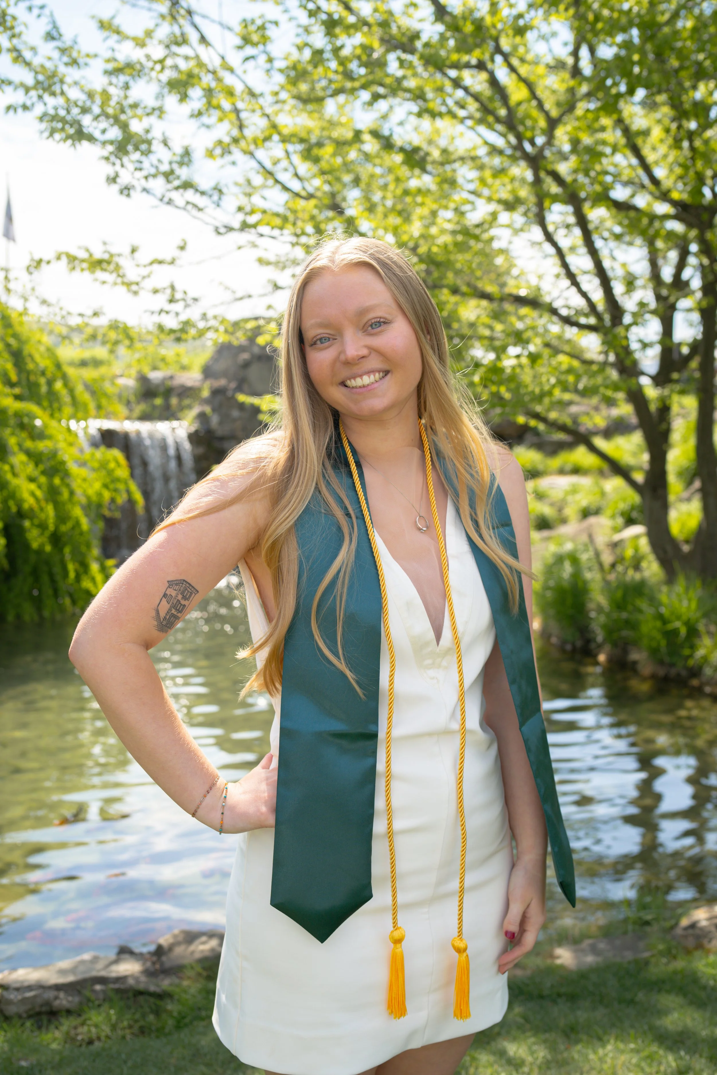 A young woman in a white dress and graduation gown with yellow tassels, standing outdoors by a river with green trees in the background, smiling at the camera.