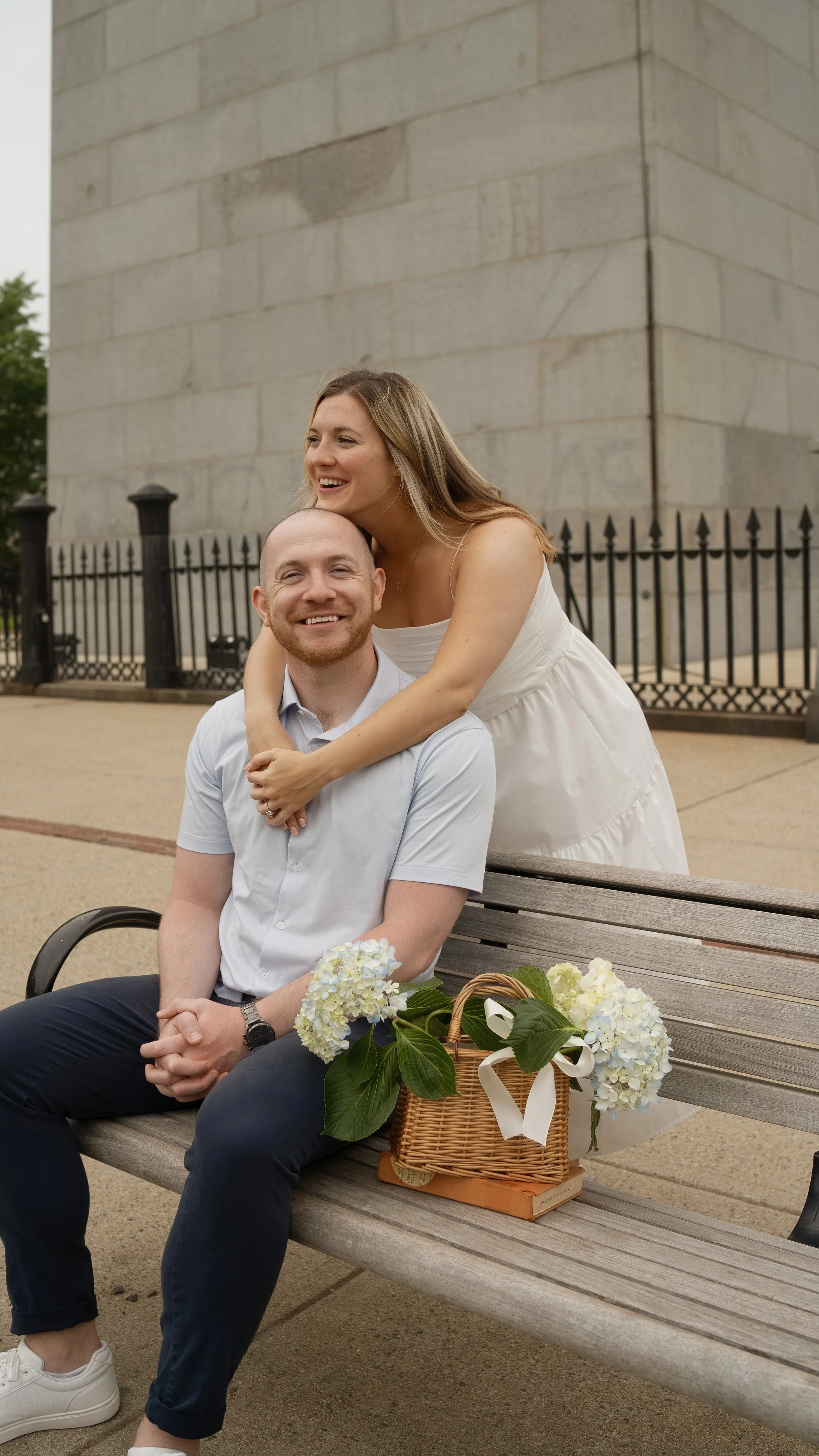 A smiling couple sitting on a bench outdoors, with a woman in a white dress leaning on the man's shoulder. There are hydrangea flowers in a basket on the bench.