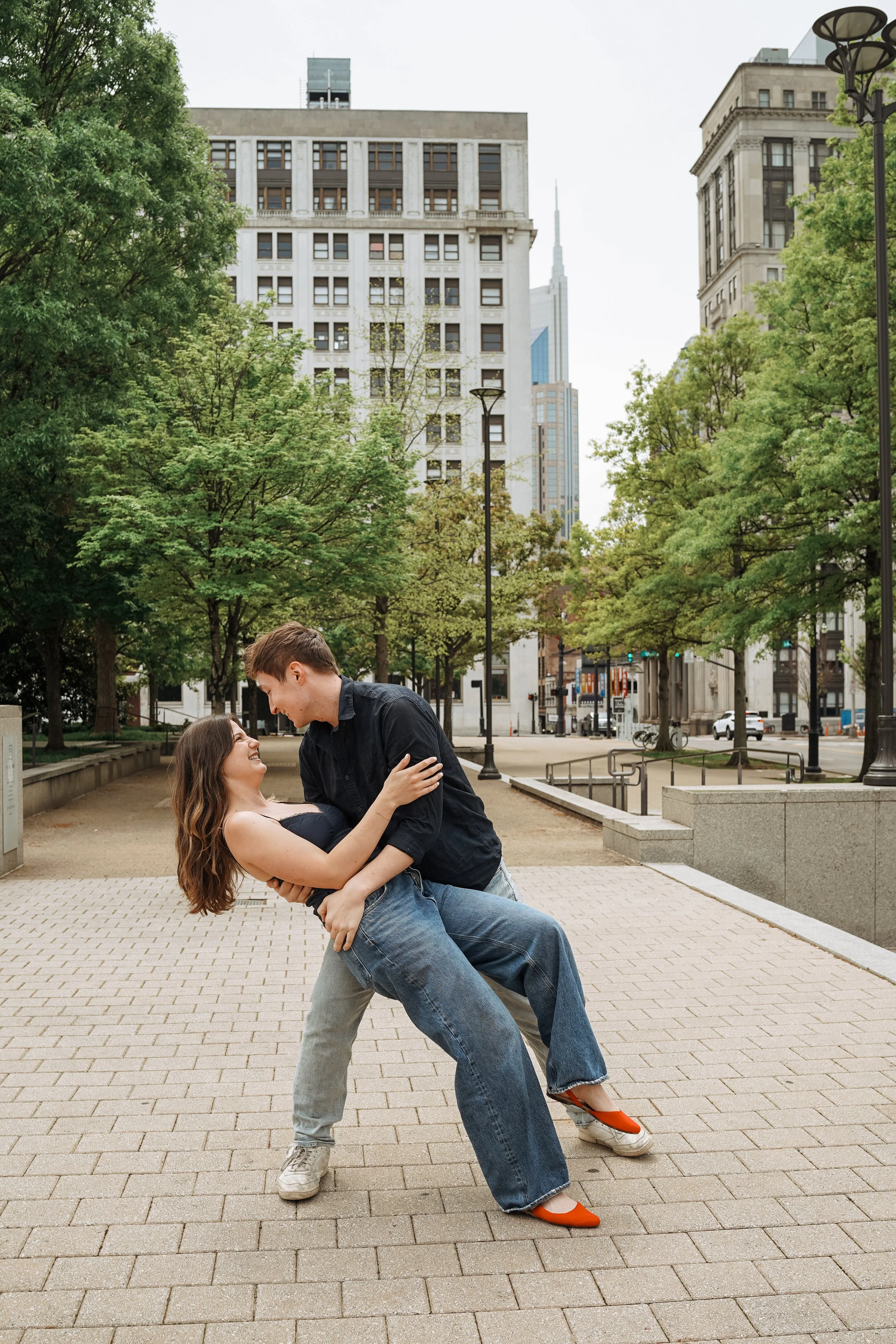 A young man and woman are dancing together in an urban park, with the man lifting the woman as they smile at each other amidst green trees and city buildings in the background.