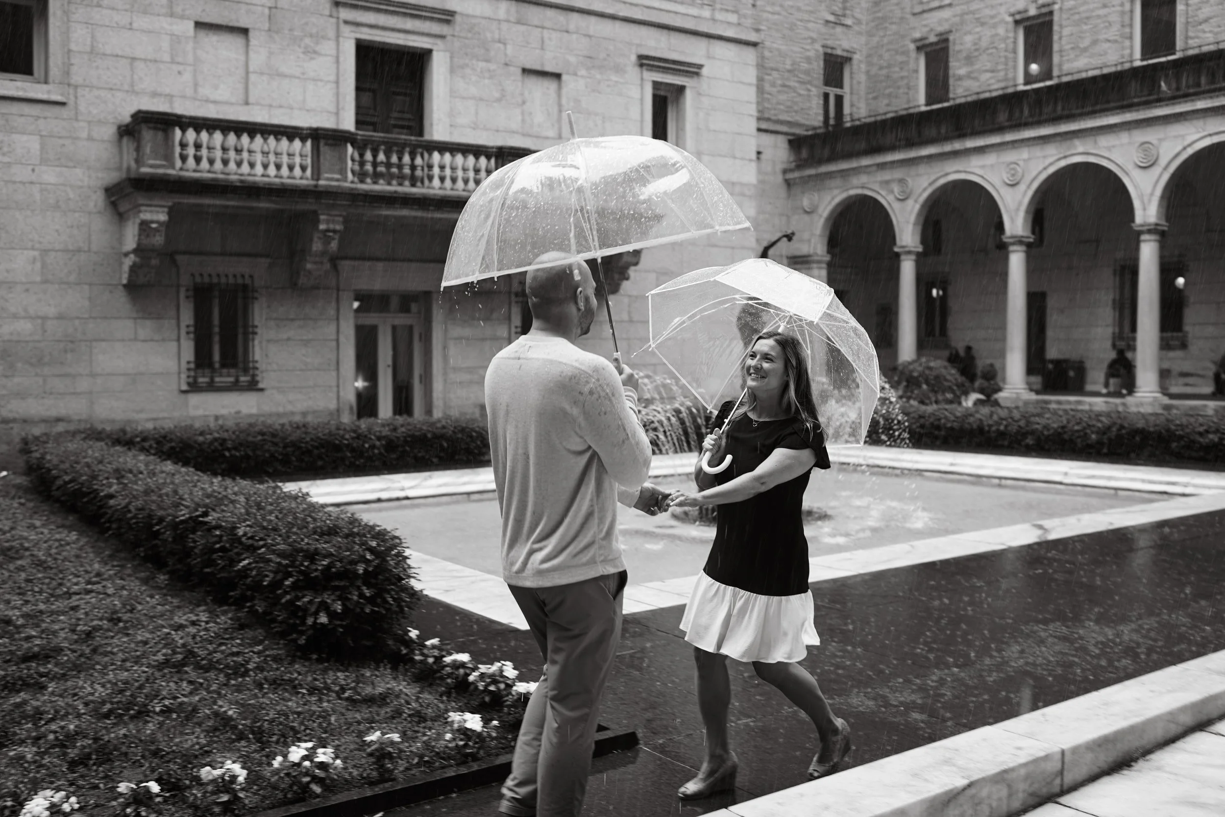 A man and woman are holding umbrellas and smiling at each other outdoors in rain. They are on a city street with historical buildings and a fountain in the background.