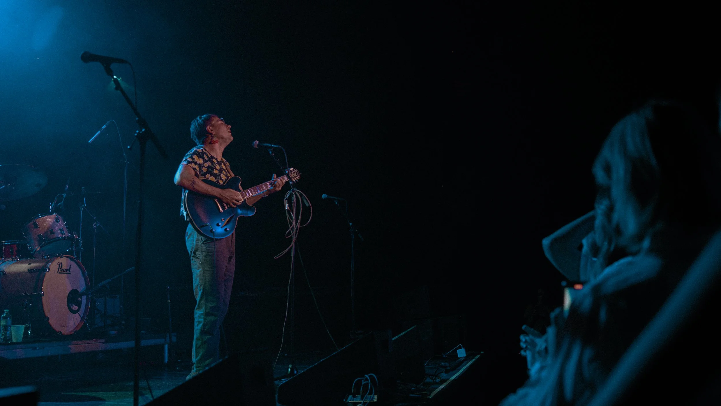 Musician playing guitar and singing on stage during a live concert, with a drum set visible in the background and audience members in the foreground.