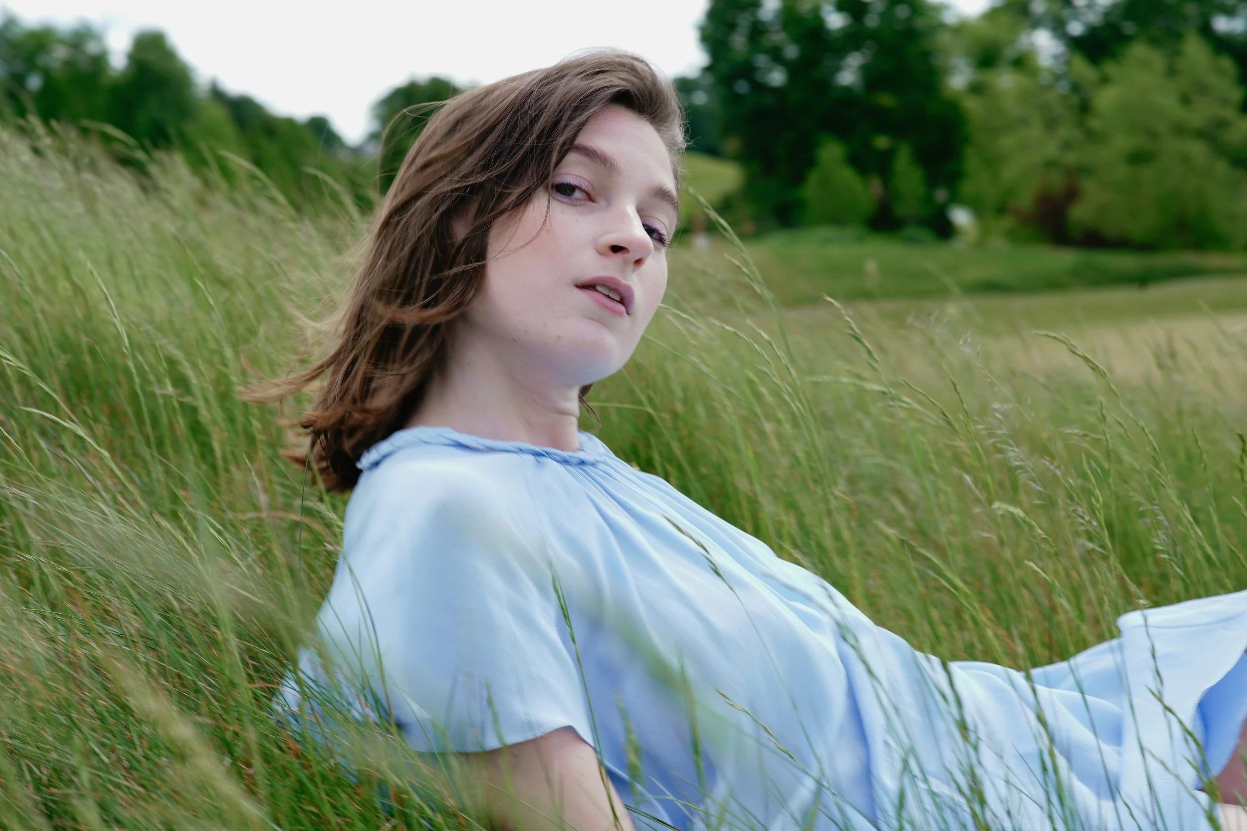 A young woman with brown hair and fair skin sitting in a grassy field with trees in the background, wearing a light blue blouse.