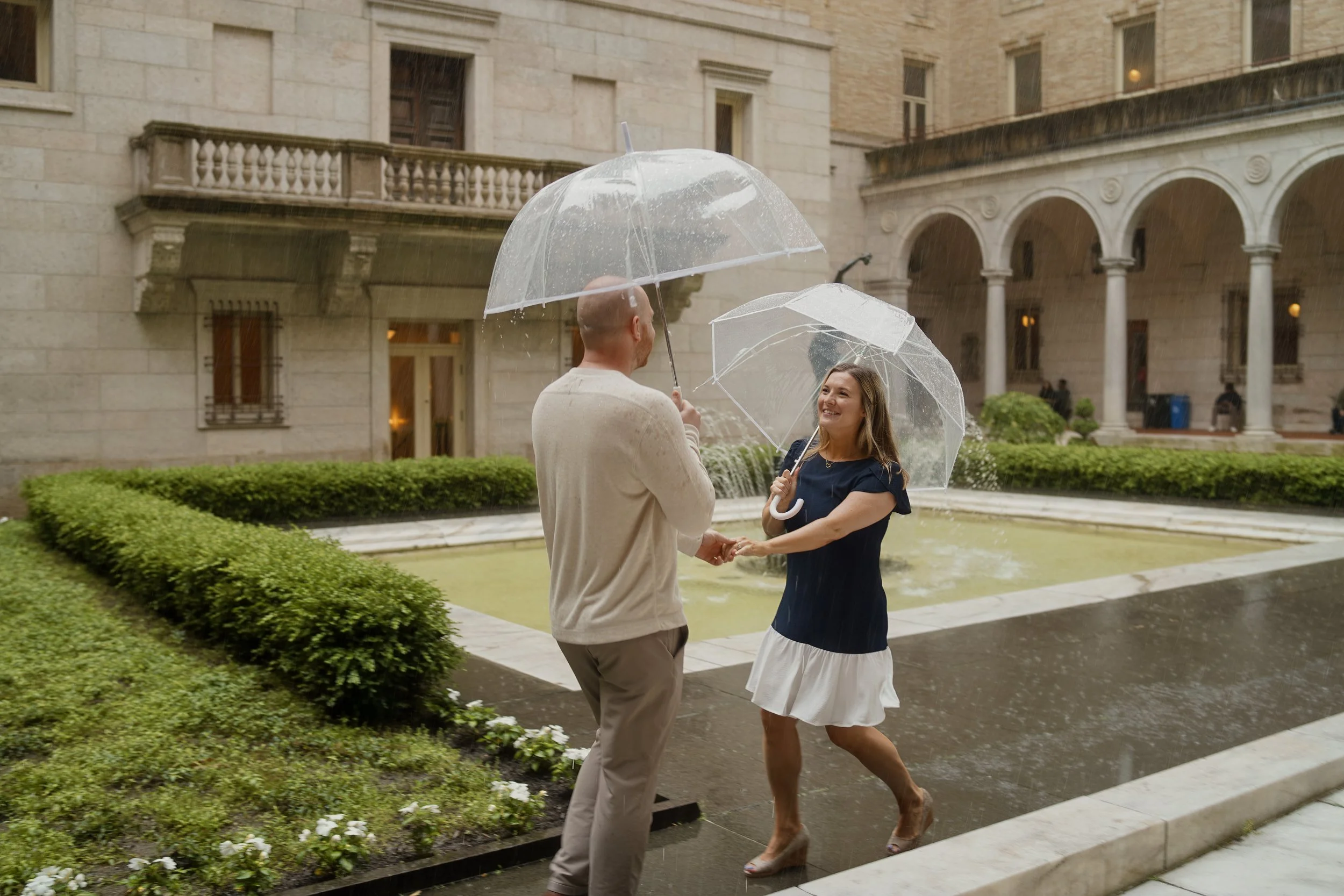 A man and woman sharing an umbrella on a rainy day in front of a stone building with arches and a fountain.