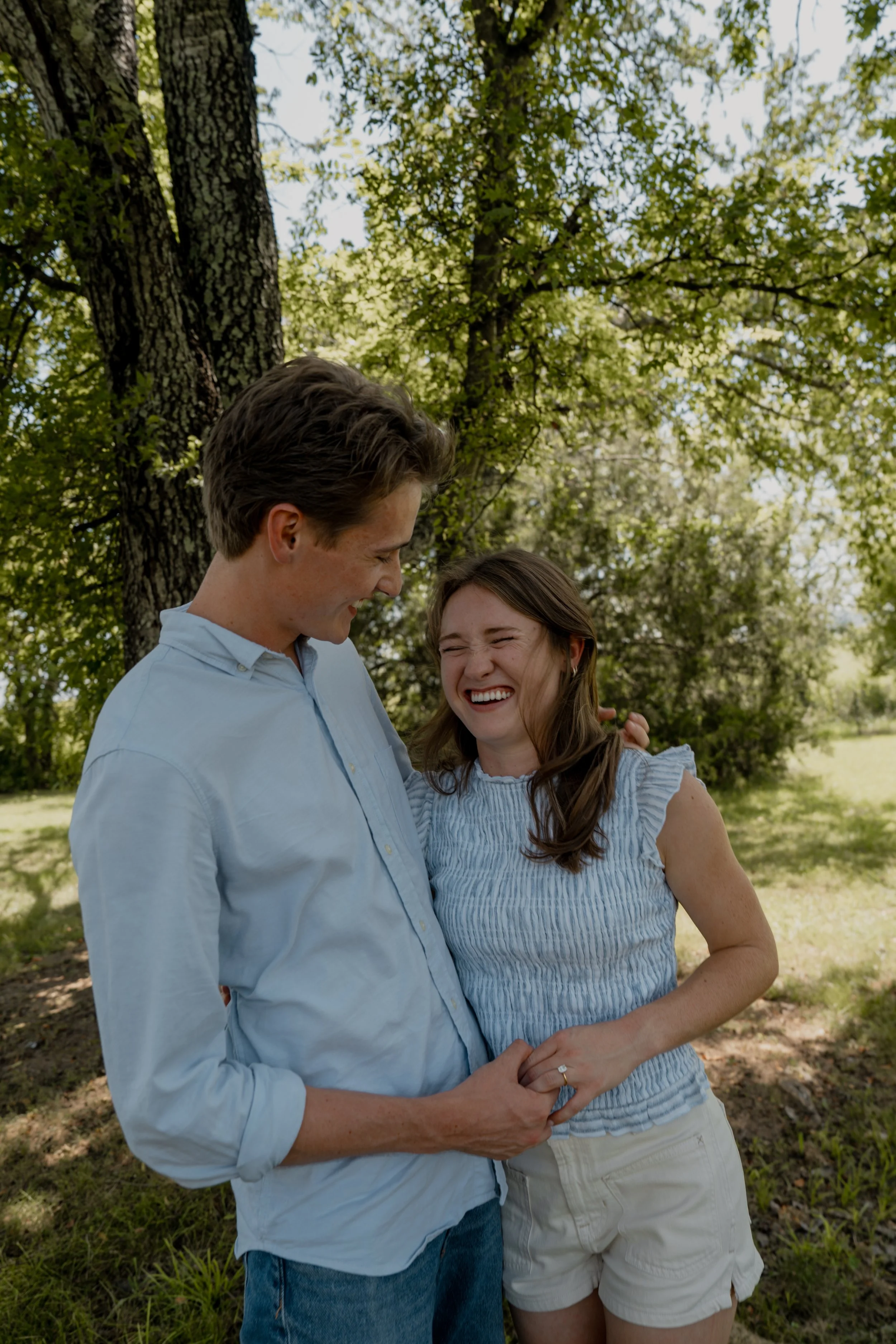 A smiling couple stands outdoors during daytime, holding hands and looking at each other affectionately. The man has short brown hair, is wearing a light blue shirt, and the woman has long brown hair, is wearing a striped sleeveless top and white sho