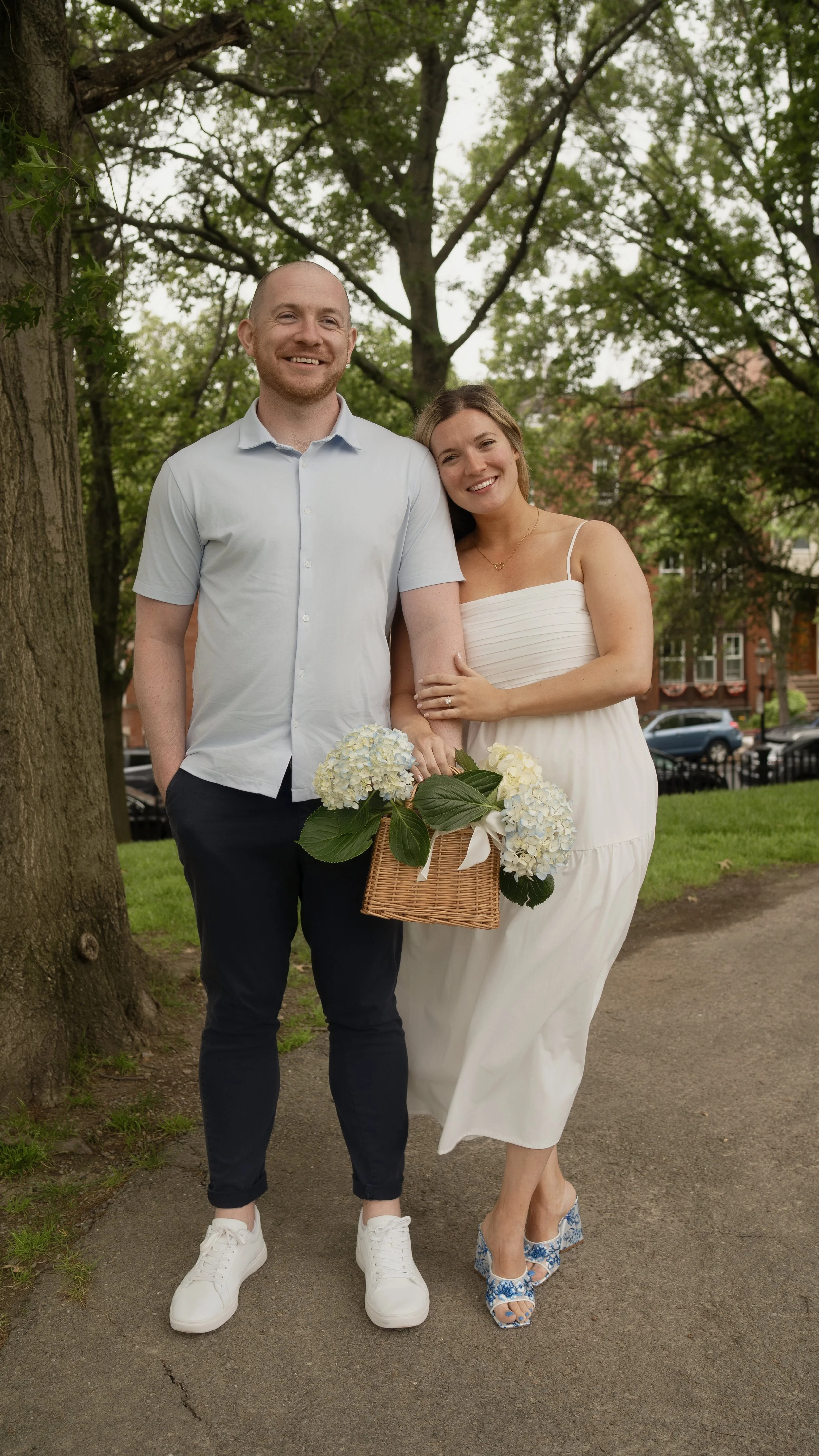 A smiling man and woman standing outdoors under a large tree, holding a basket with white and blue hydrangea flowers, with houses and parked cars visible in the background.