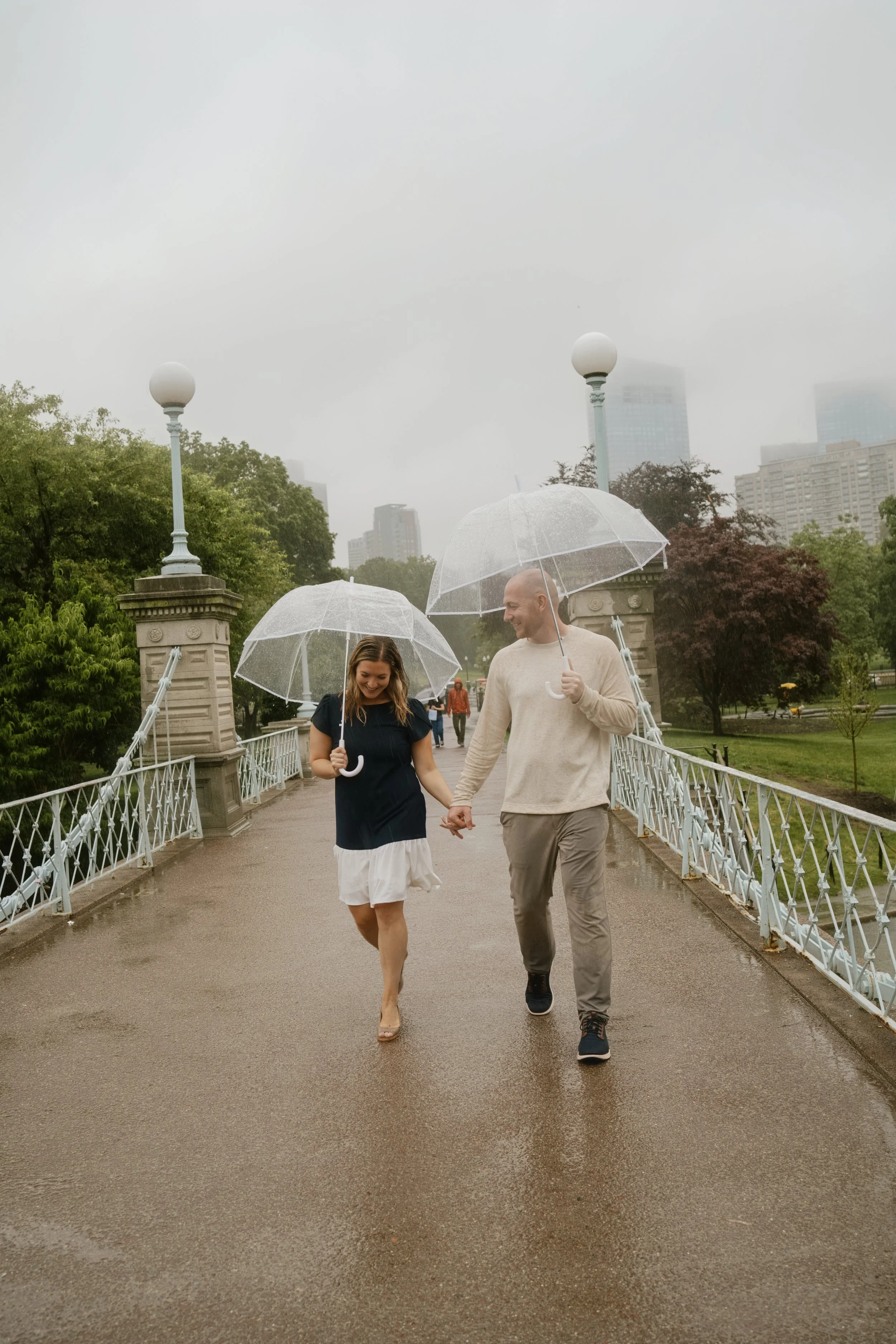 A couple walking on a bridge with white railings during a rainy day, holding clear umbrellas, surrounded by green trees with a city skyline in the background.