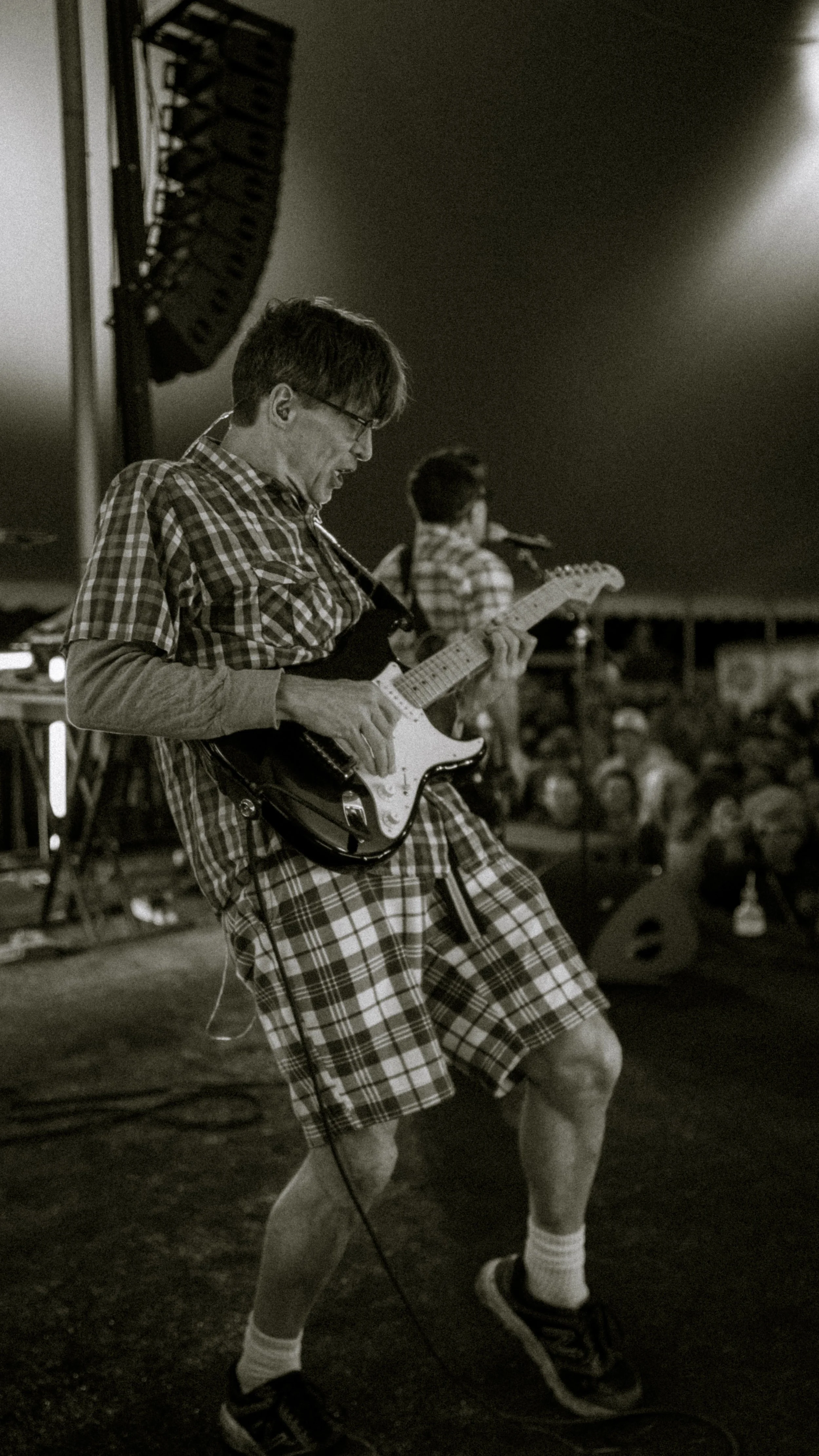 A young man wearing plaid shorts, a plaid shirt, sneakers, and glasses playing an electric guitar on stage during a concert. He has a joyful expression and is dancing with his eyes closed.
