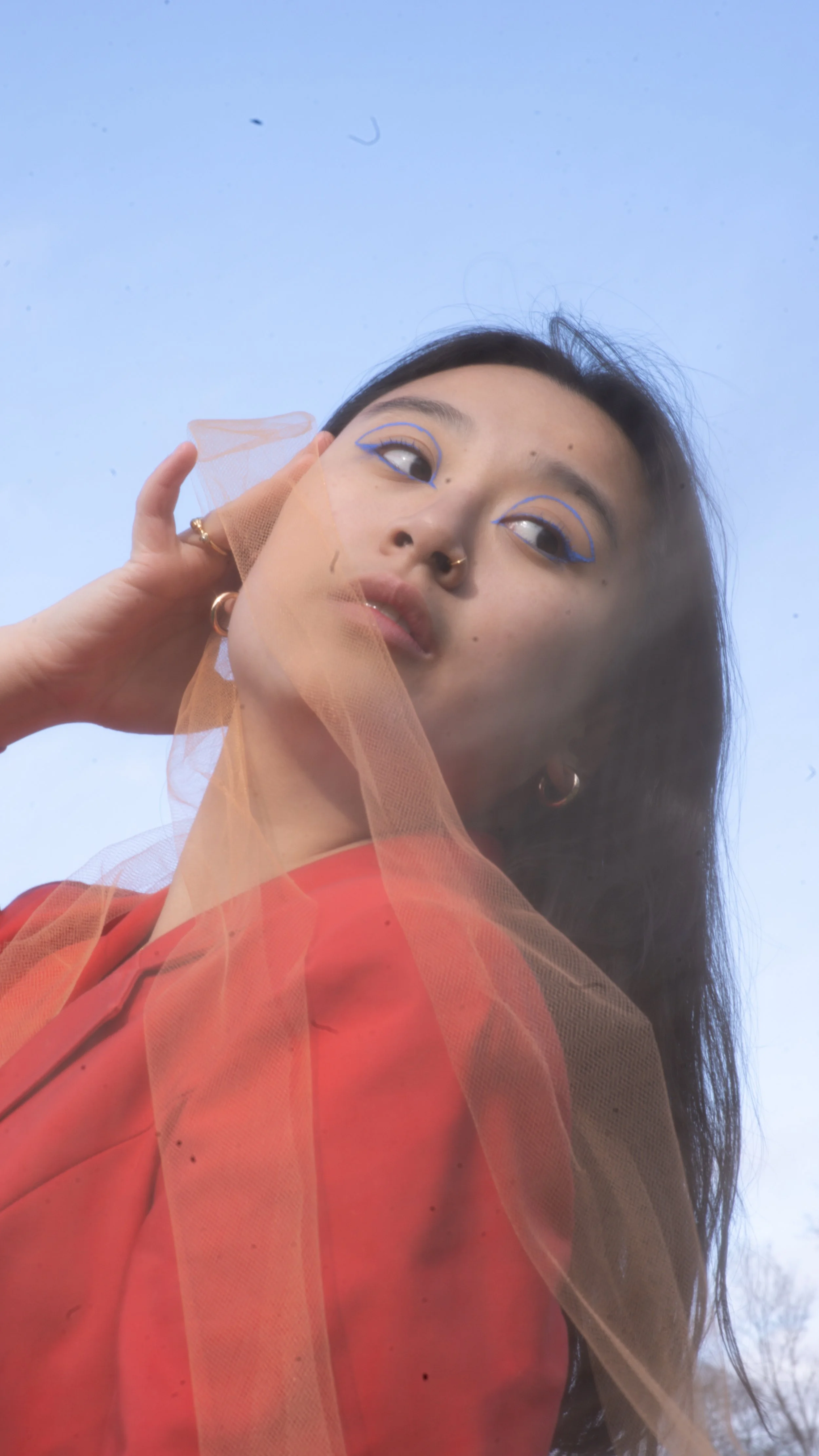 A woman with dark hair and multiple facial piercings, wearing a red blazer with an orange tulle overlay, looking upward against a light blue sky.