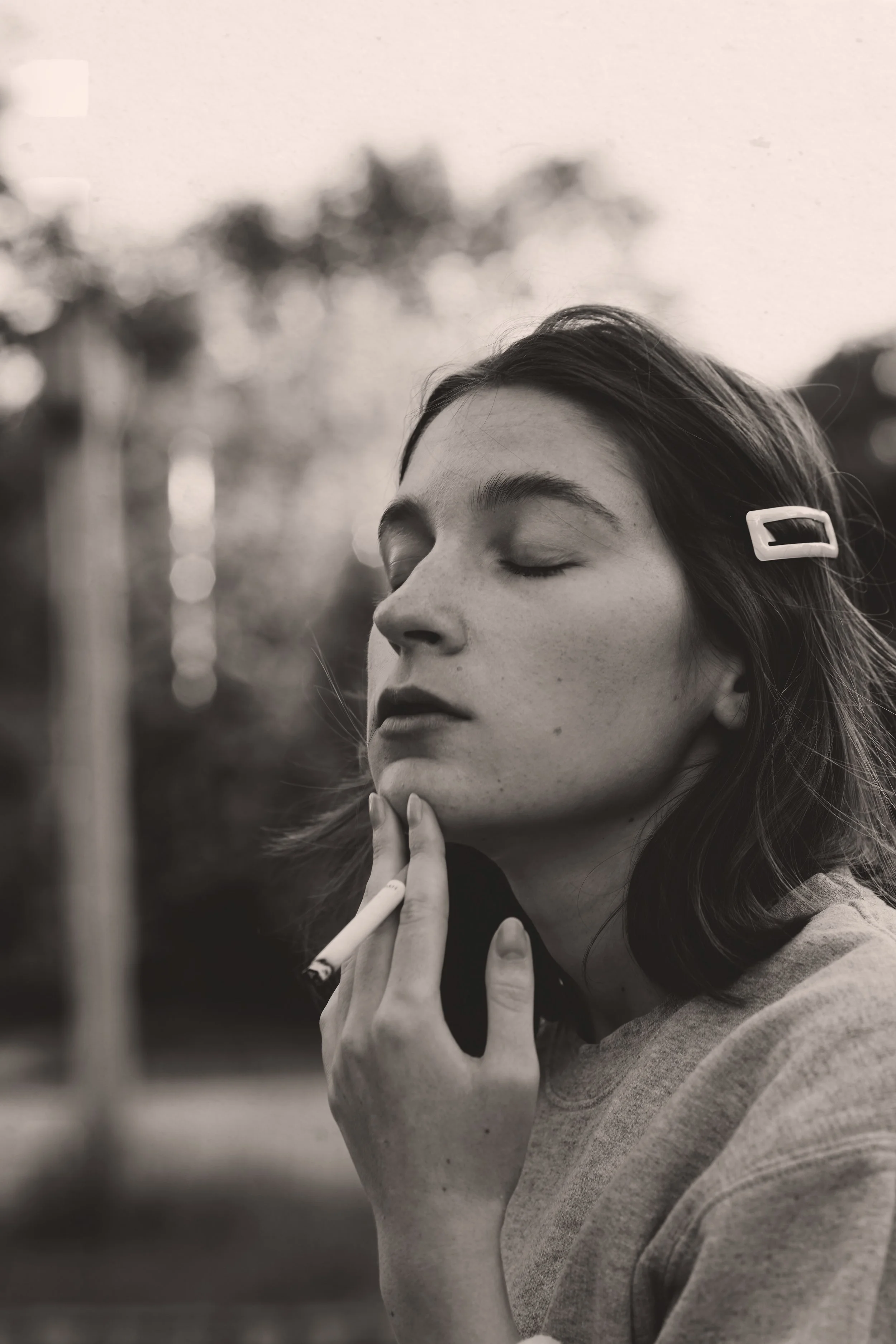 Black and white photo of a woman with closed eyes, holding a cigarette near her neck, with her hair clipped back with a white hair clip, and wearing a light sweatshirt, outdoors with blurred trees in the background.