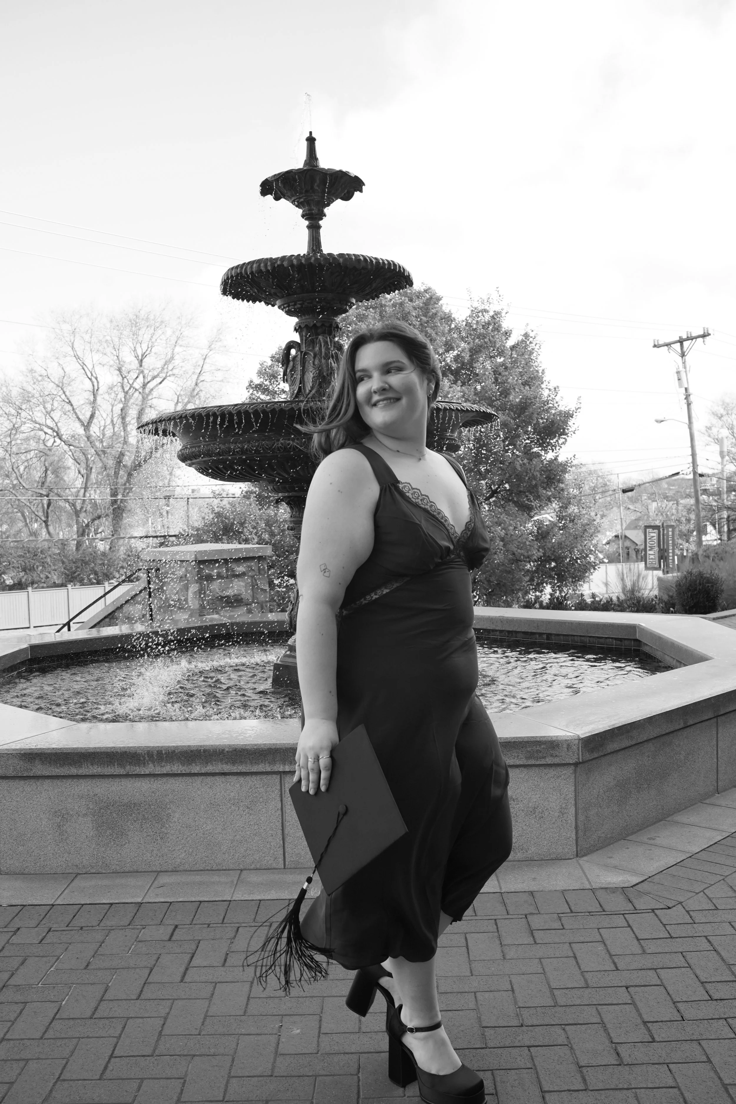 A woman in a black dress and high heels stands next to a three-tiered fountain with water spouts, holding a small clutch purse, and smiling at an outdoor location with trees, power lines, and a sign in the background.