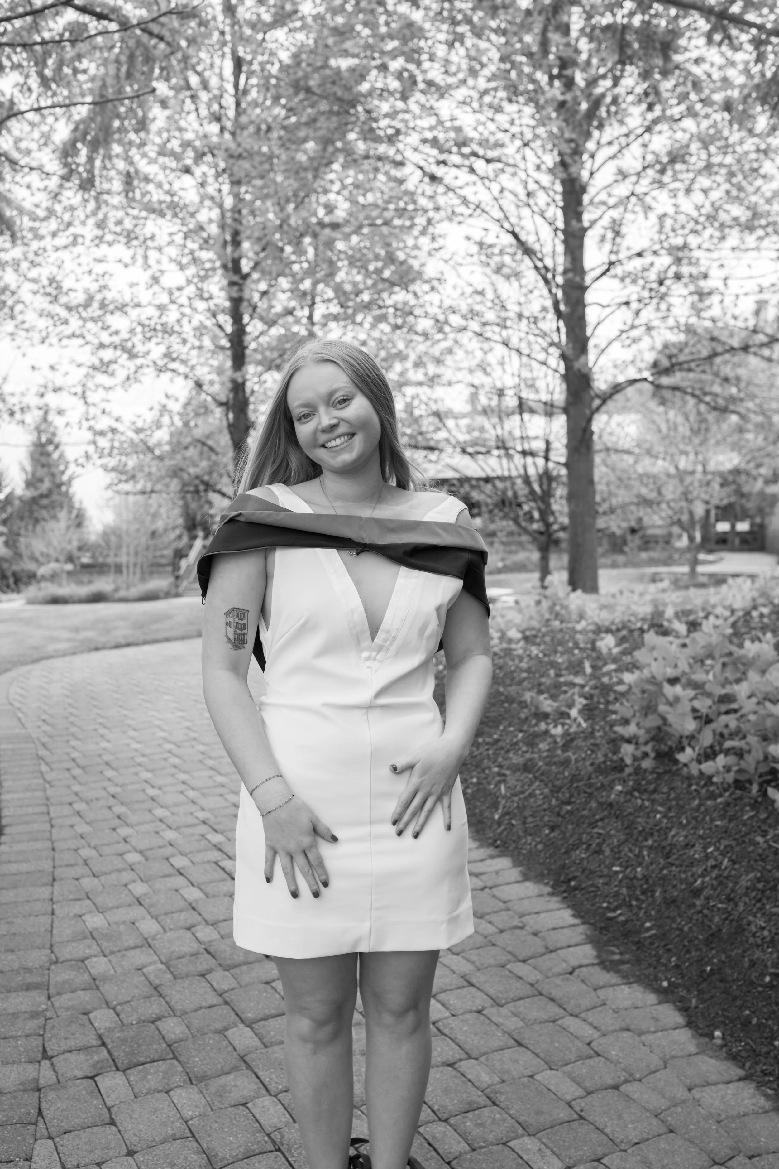 A woman in a sleeveless dress with a cape over her shoulders standing outdoors on a brick pathway in a park with trees and foliage in the background.