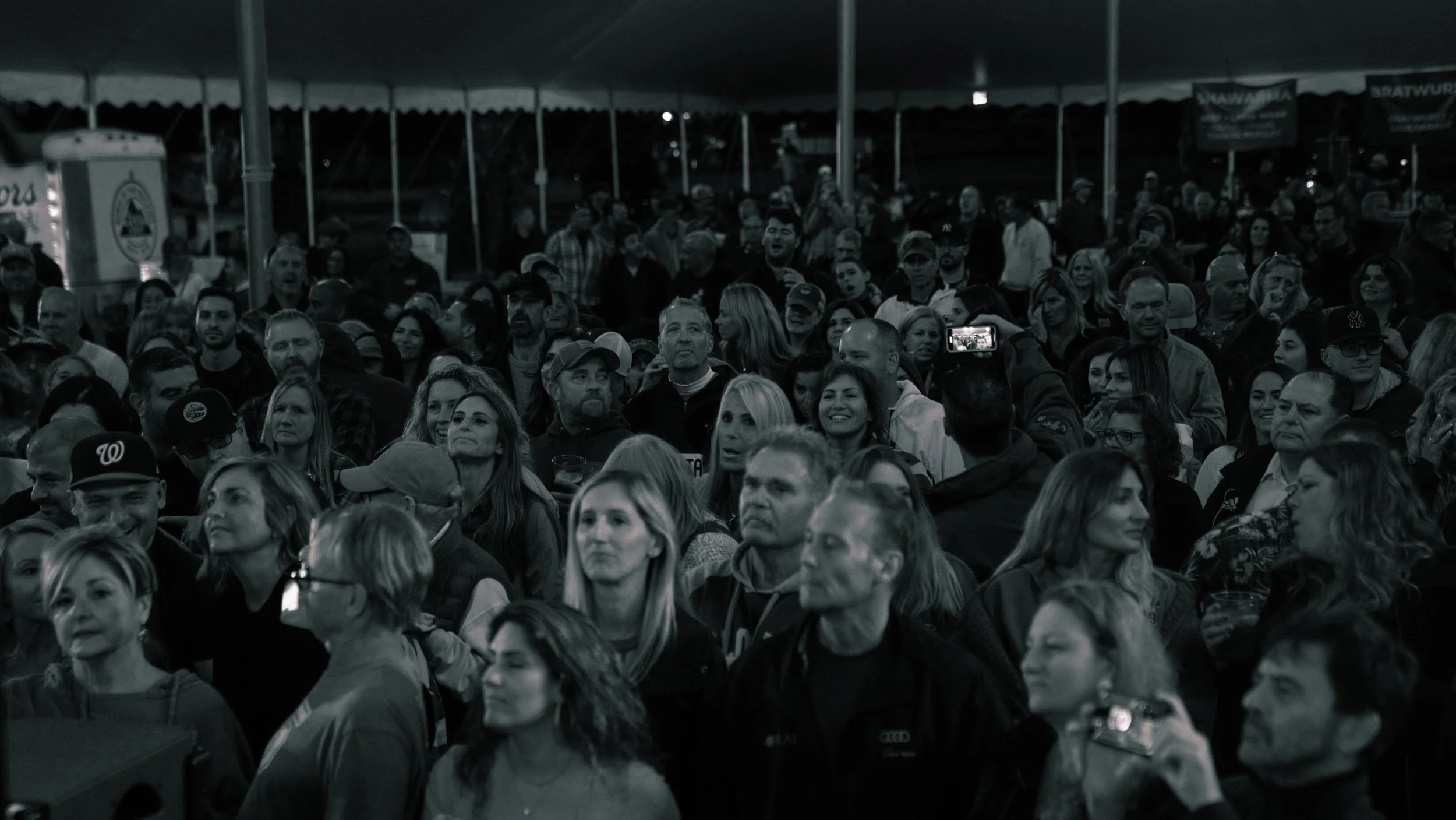 Crowd of people at an outdoor event, many facing forward, some smiling, some talking, under a large tent at night.