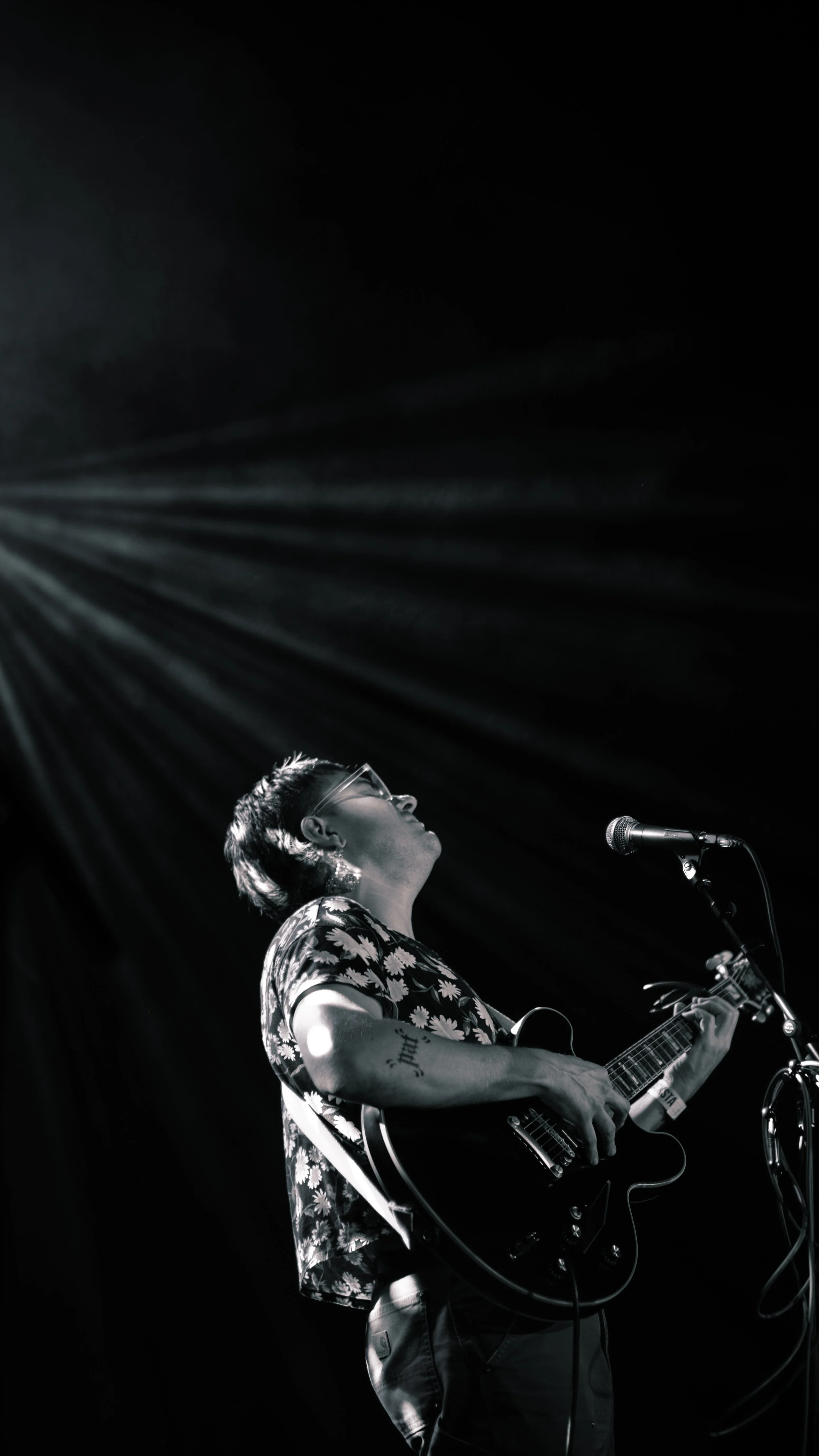 A person wearing glasses and a floral shirt is playing an acoustic guitar and singing into a microphone on stage with a dark background and stage lights shining down.