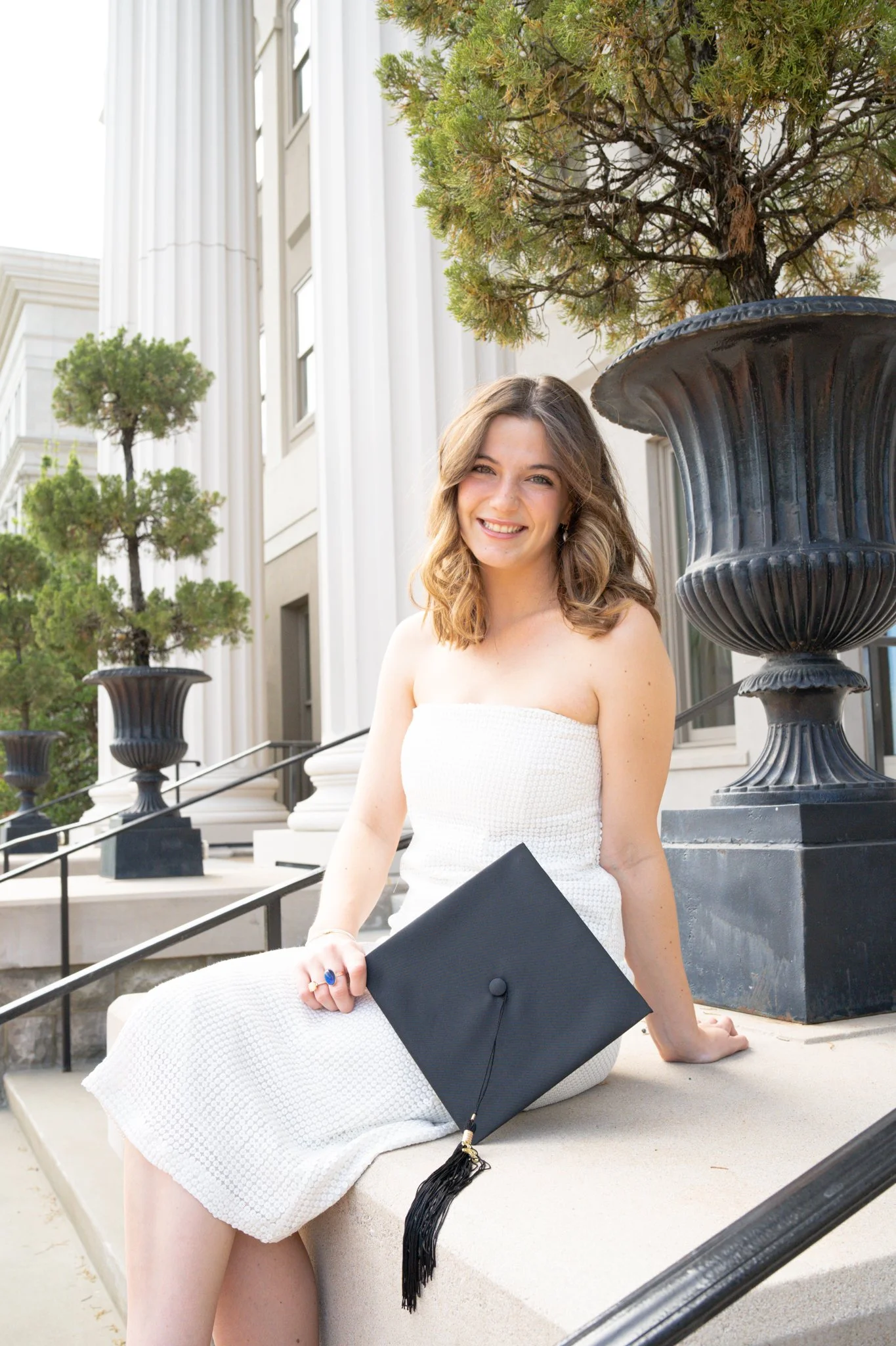 A young woman with wavy brown hair, smiling, wearing a strapless white dress, sitting on steps outside a building with white columns, holding a black diploma cover and wearing a graduation cap, with large planters and trees in the background.