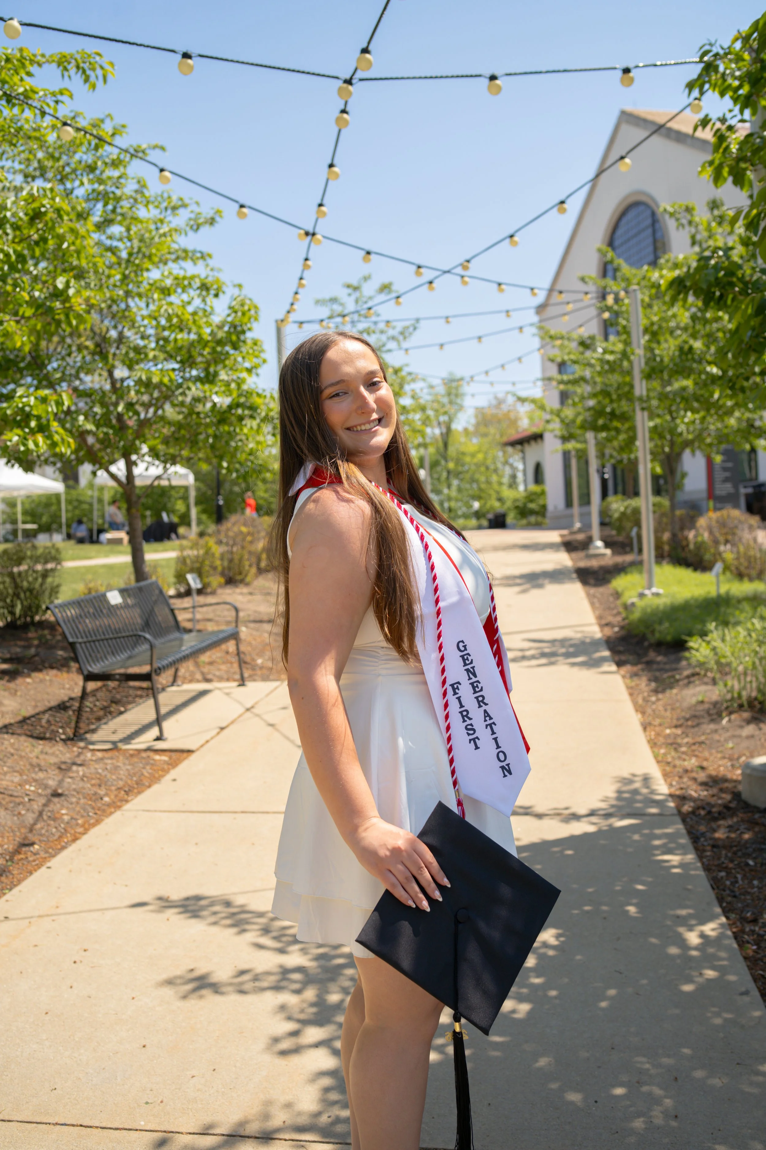 A young woman in an off-white dress standing outdoors, smiling, holding a black diploma cover, and wearing a white sash with blue text that reads 'First Generation Graduation' in front of a church and decorated with string lights on a blue sky day.