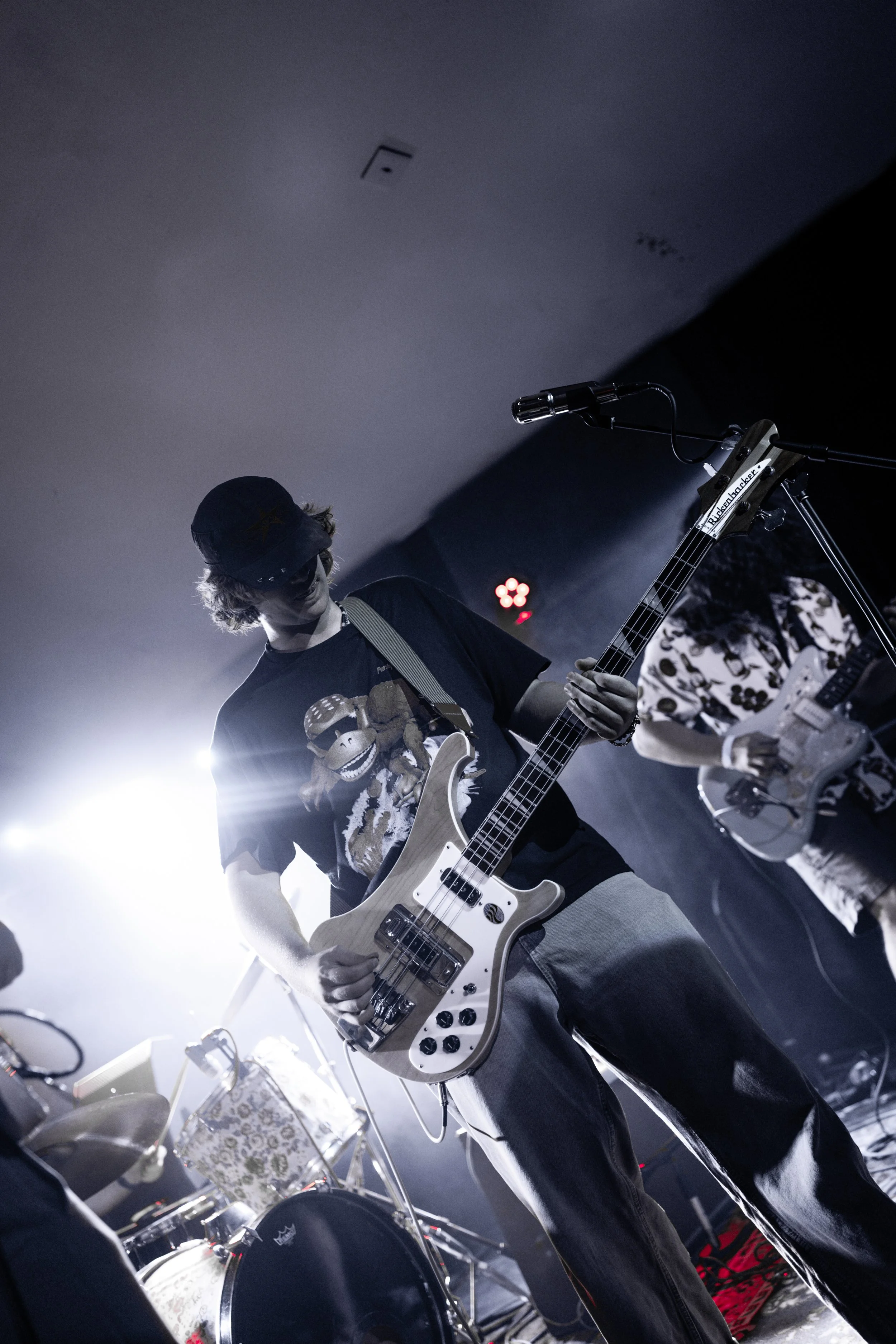 A musician playing an electric guitar on stage, wearing a cap and a black graphic t-shirt, with drums and equipment in the background, illuminated by stage lighting.