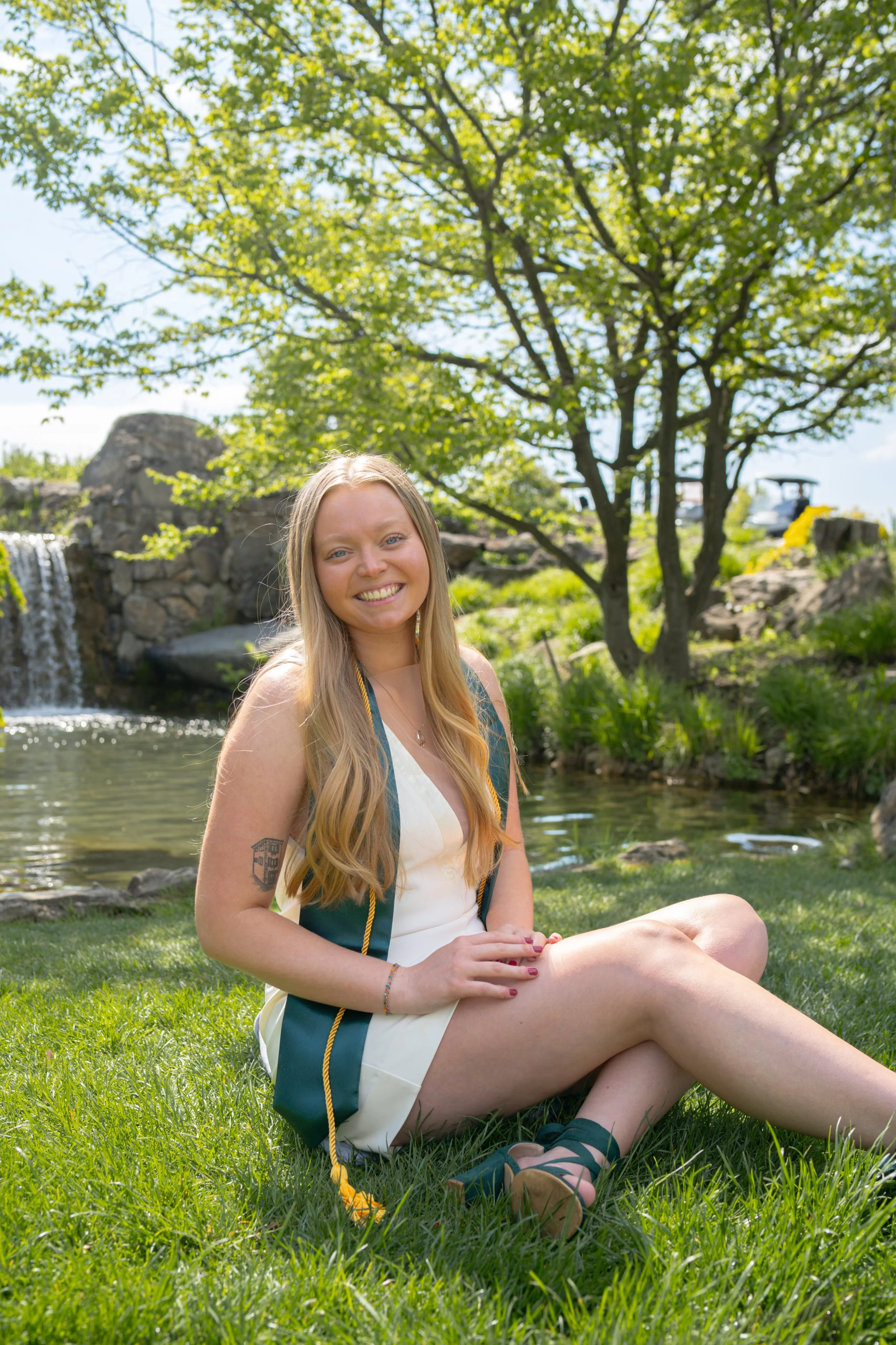 A young woman with long blonde hair and tattoos on her arm is sitting on the grass by a pond with trees and rocks in the background, smiling at the camera.