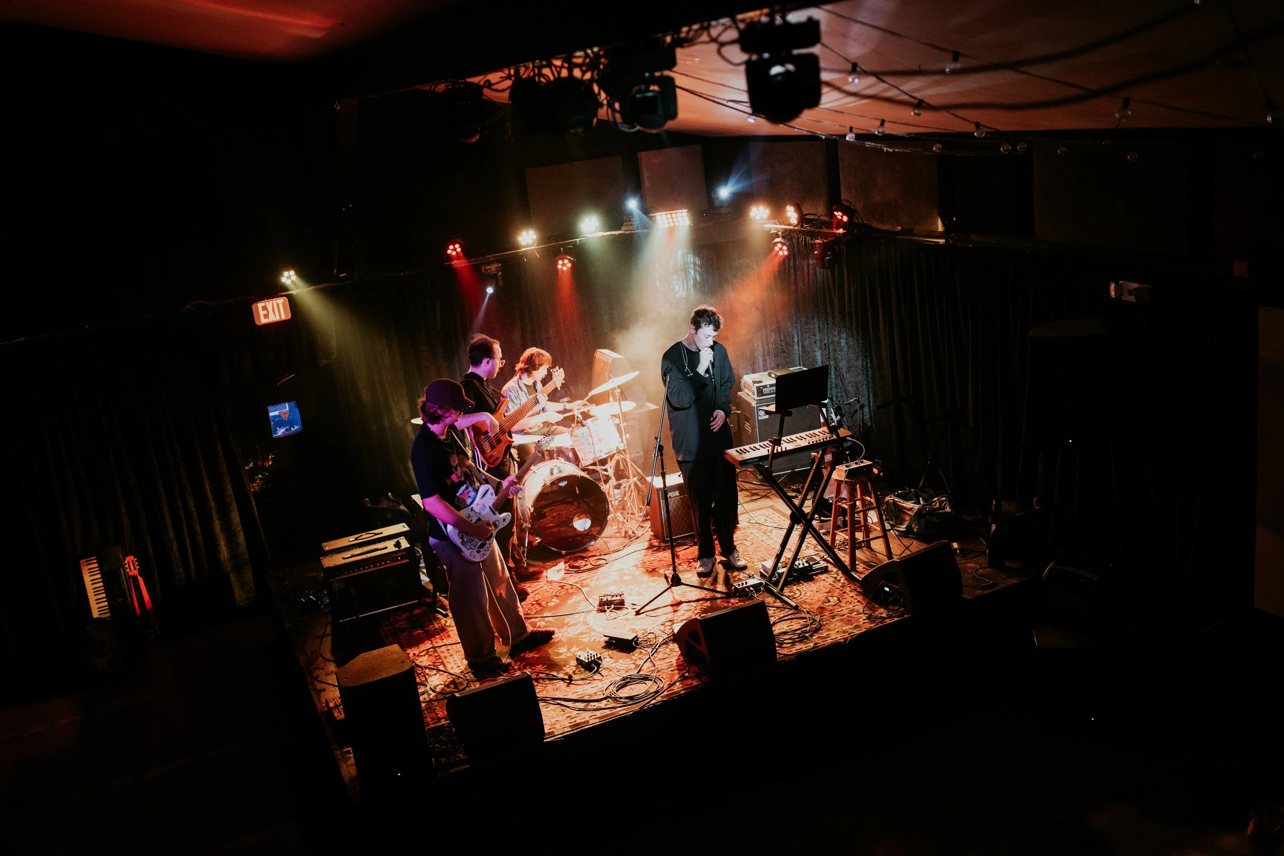 A band performing on a stage with colorful lighting, including a drummer, a keyboardist, a guitarist, and a singer, in a venue with dark curtains and an exit sign.