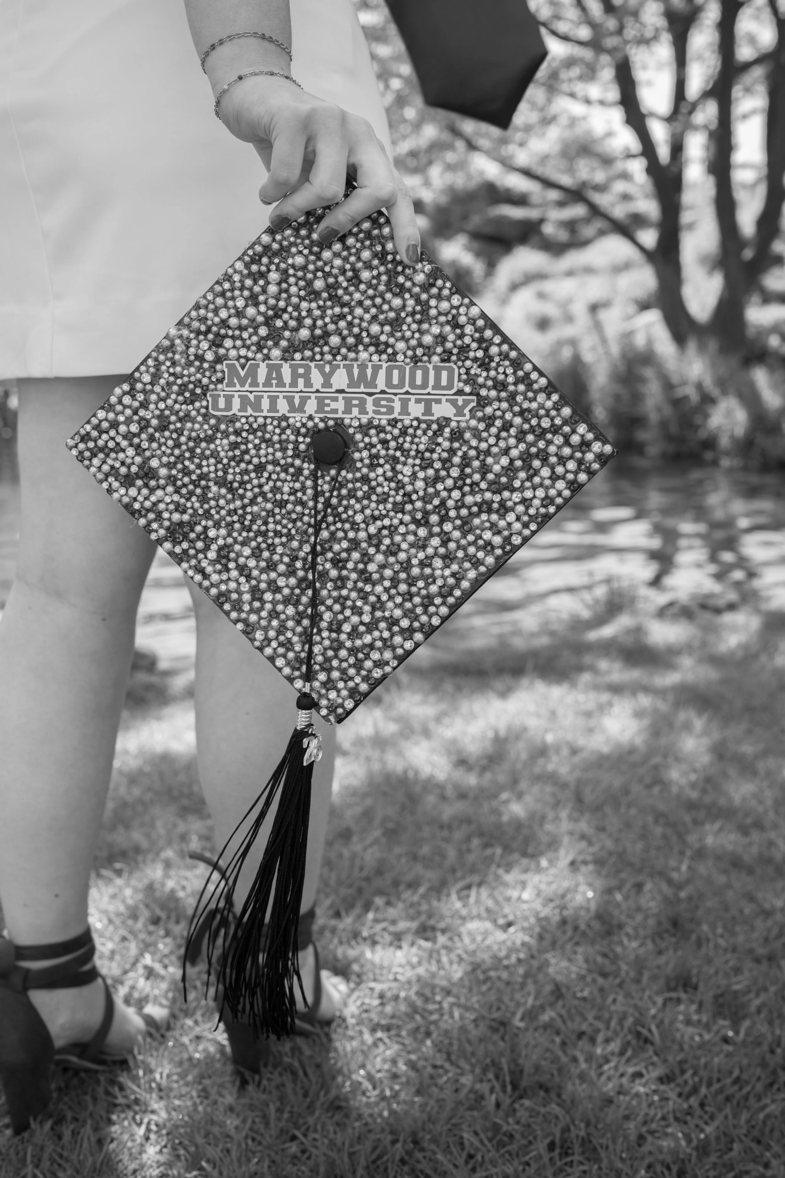 Close-up of a person's hand holding a decorated cap with beads and a sticker reading "Marywood University" during a graduation ceremony outdoors.