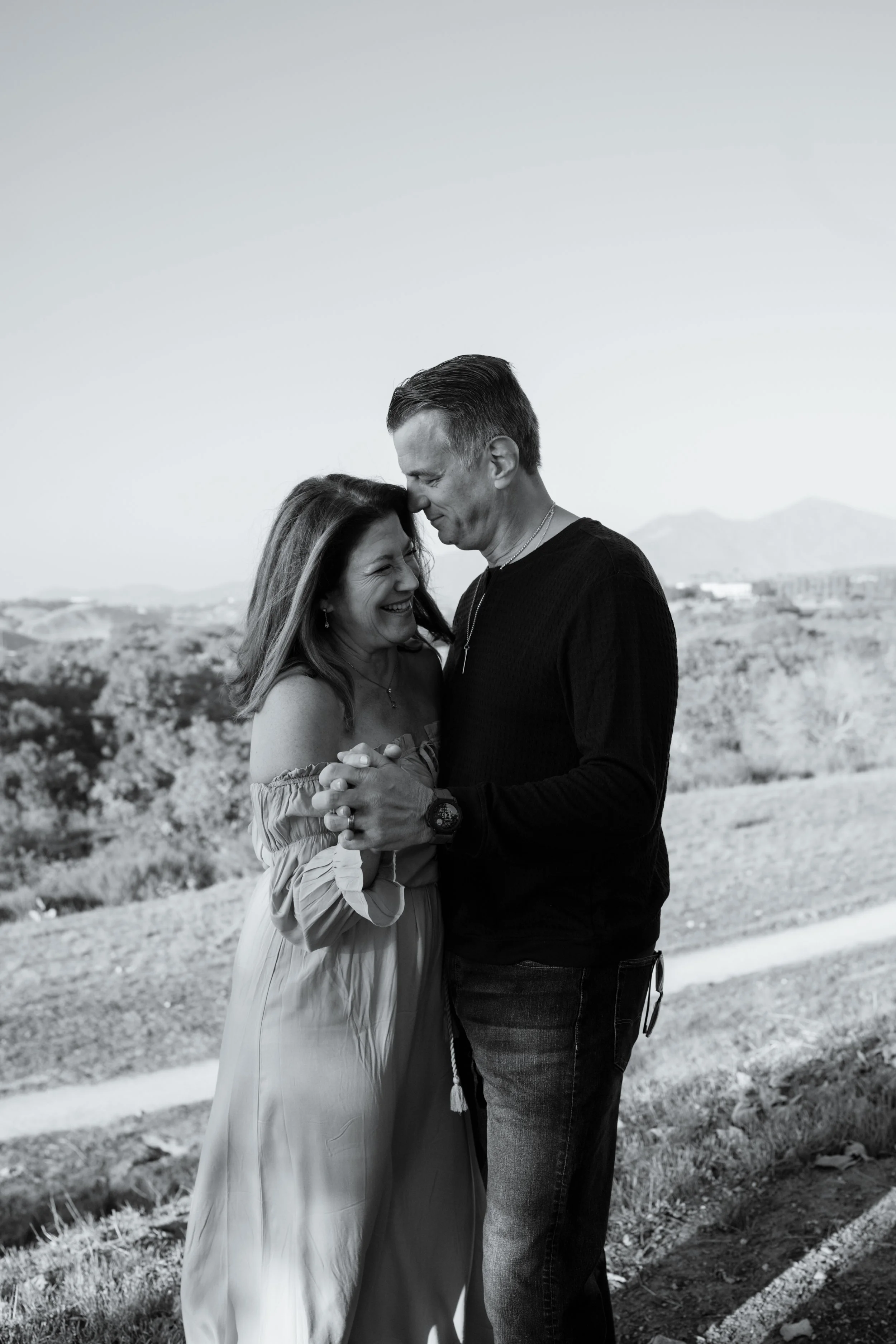 A black-and-white photo of a smiling woman and man holding hands and looking at each other outdoors, with mountains and a landscape in the background.