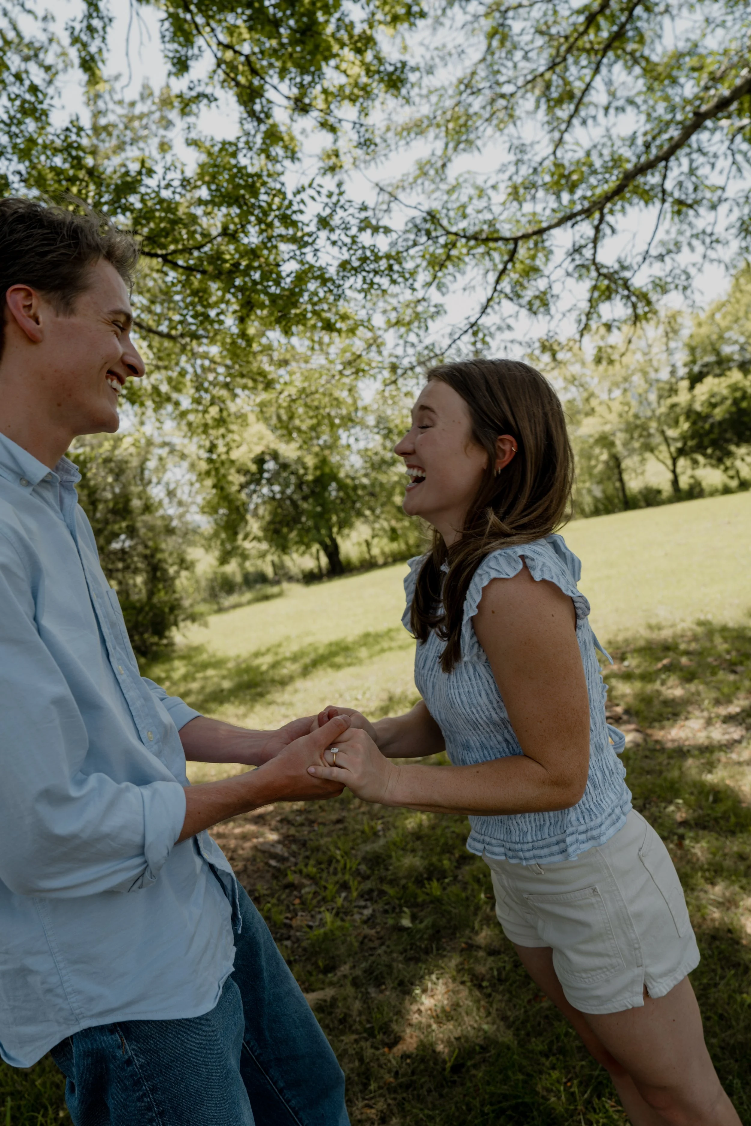 A couple holding hands and smiling at each other outdoors in a park with green trees and grass.