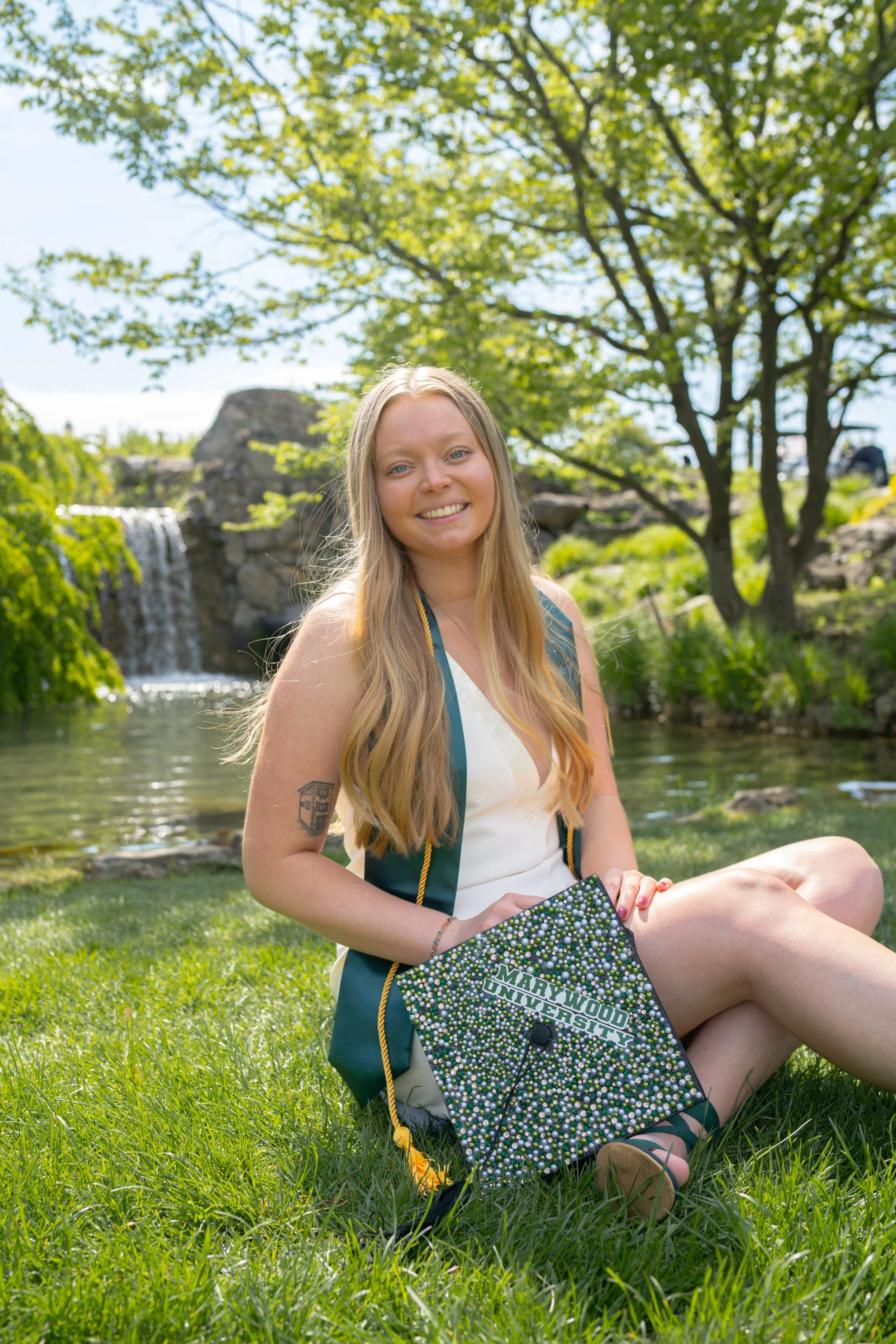 A young woman with long blonde hair, sitting on green grass near a small waterfall and river, holding a decorated graduation cap that reads 'Marywood University.' She is smiling and wearing a blue and white graduation gown outdoors on a sunny day wit